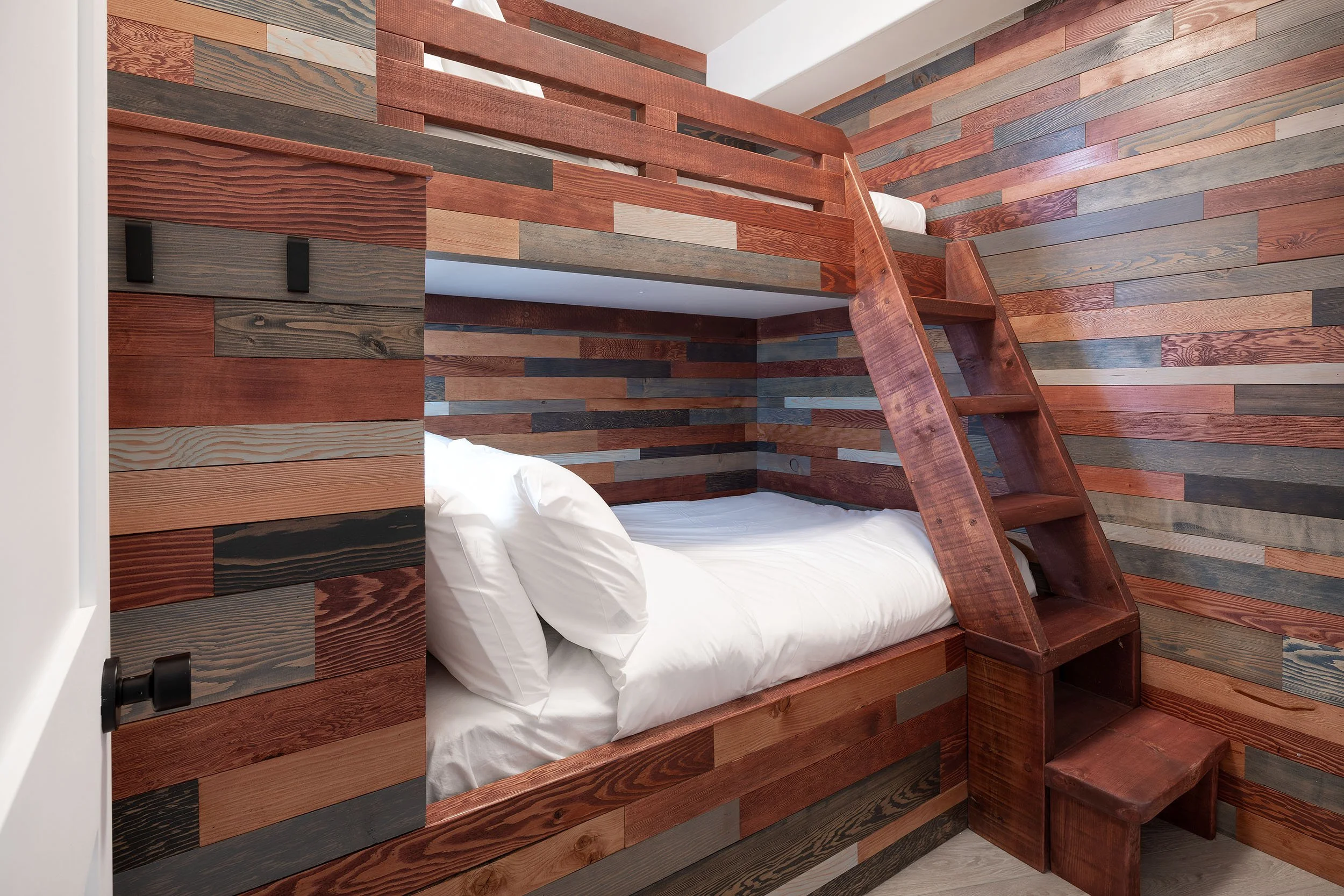 A bedroom with walls and a bedframe made from multicolored wood planks, a white bed with white pillows, and a wooden ladder.