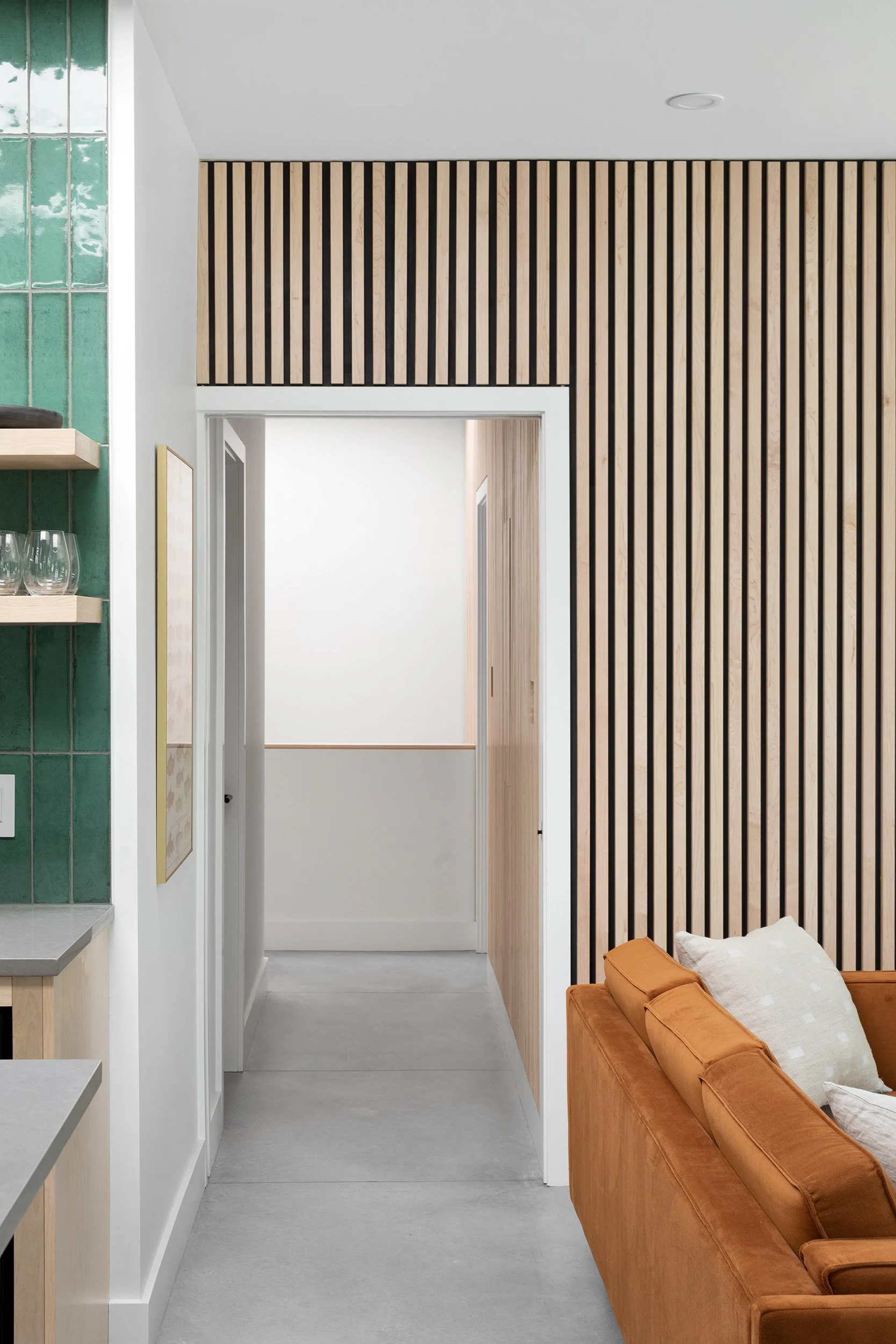 Interior of a modern living space with a wooden slat accent wall, a white doorway, a part of a kitchen with floating shelves and glassware, and a burnt orange sofa with cushions.