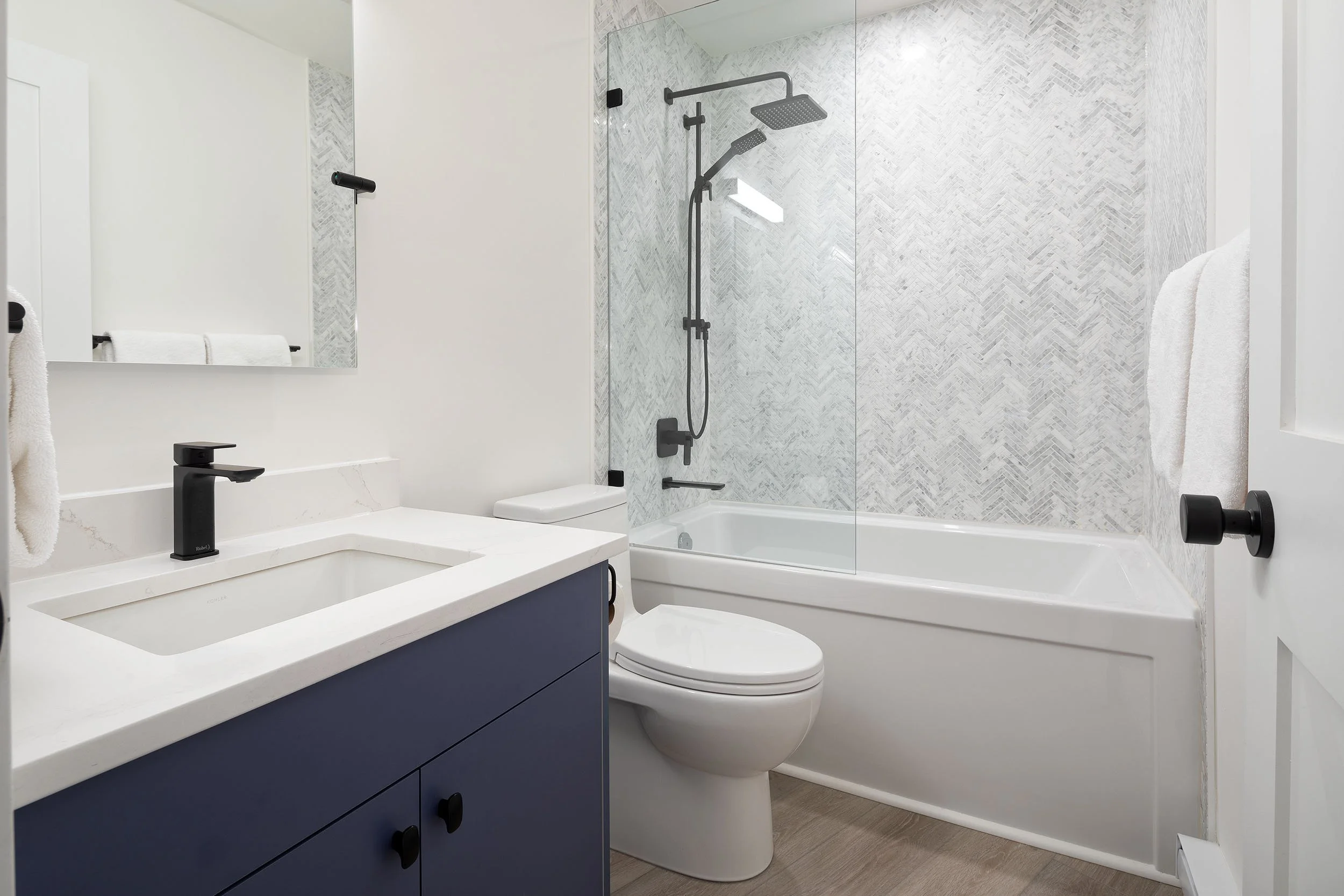Modern bathroom with white walls, a navy blue vanity with black handles and a white countertop, a rectangular sink, black faucet, and a large mirror. There is a toilet and a glass-enclosed shower with a rain showerhead and a handheld showerhead, whit