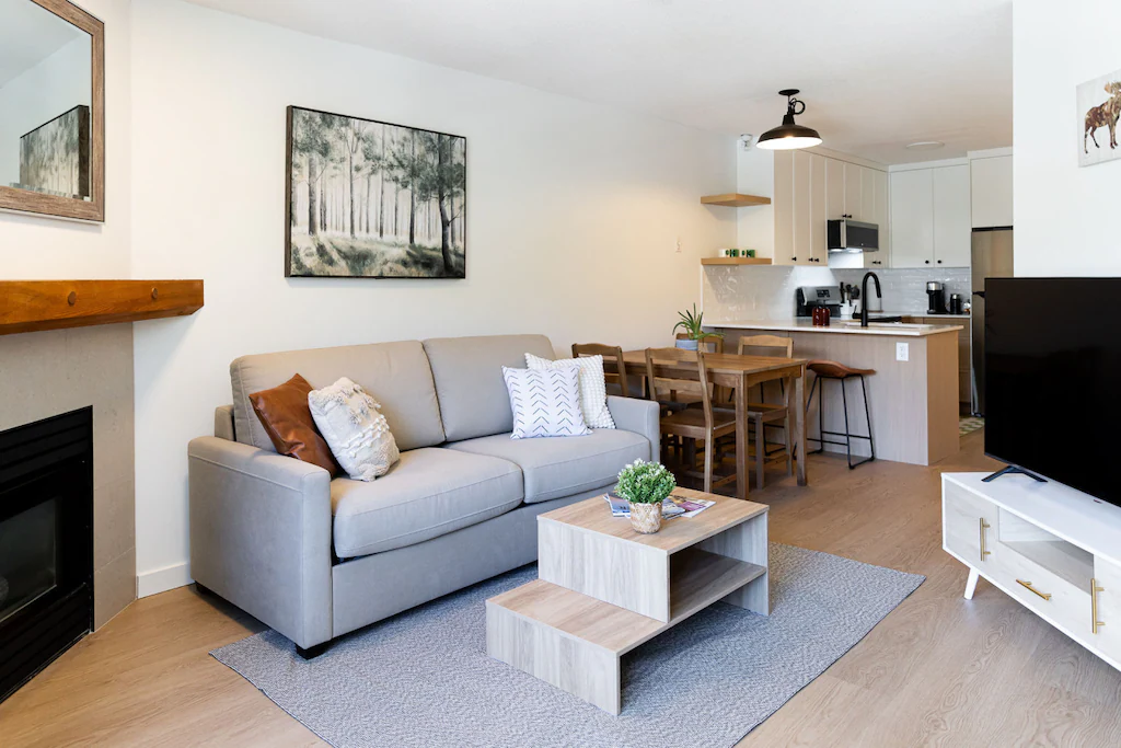 Living room with beige sofa, wooden coffee table, gray area rug, white TV stand, flatscreen TV, fireplace, framed art on white walls, open kitchen with white cabinets, wooden dining table with chairs, and kitchen appliances.