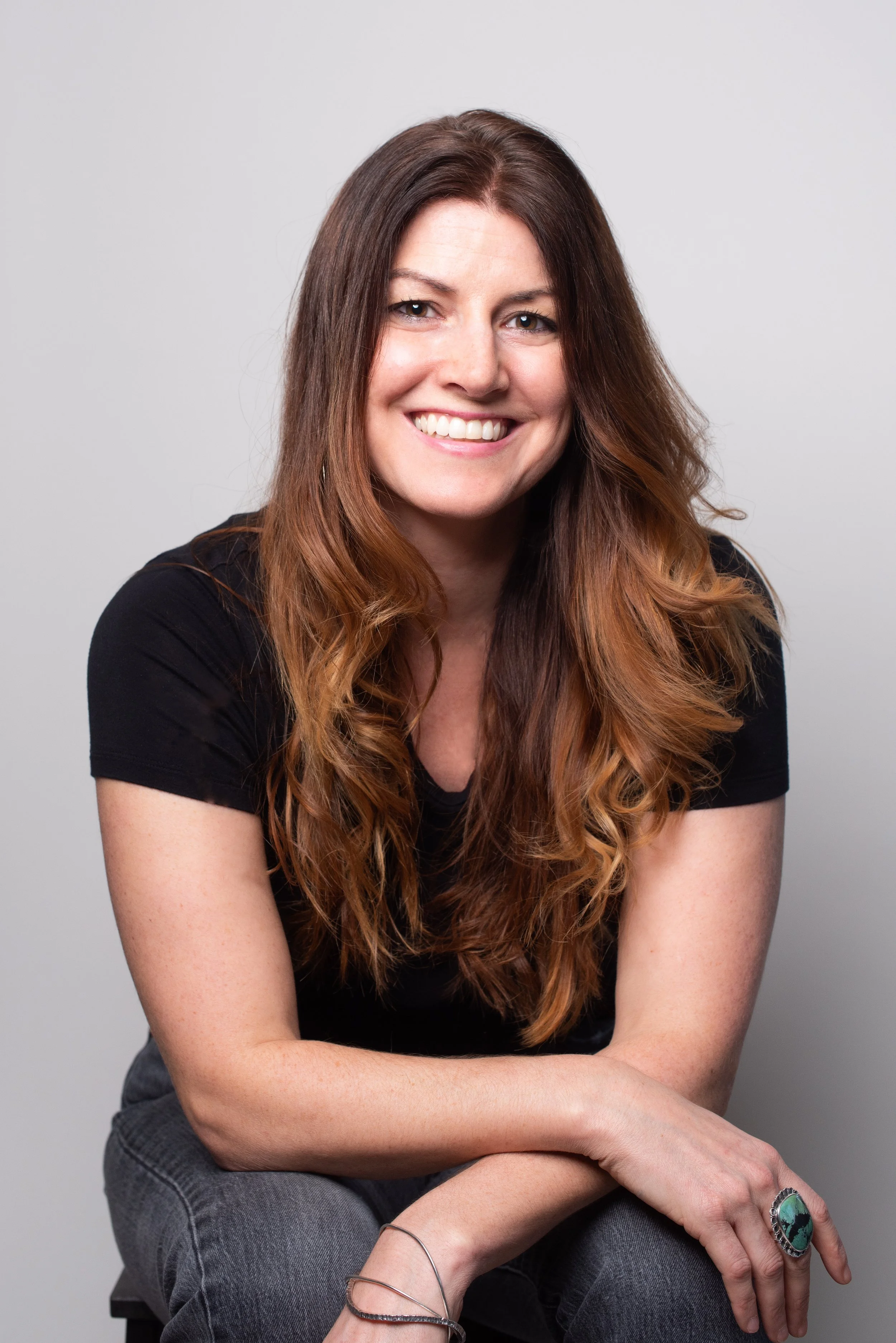 Portrait of a woman with long, wavy brown hair, smiling, wearing a black shirt, gray jeans, and jewelry, sitting against a plain light background.