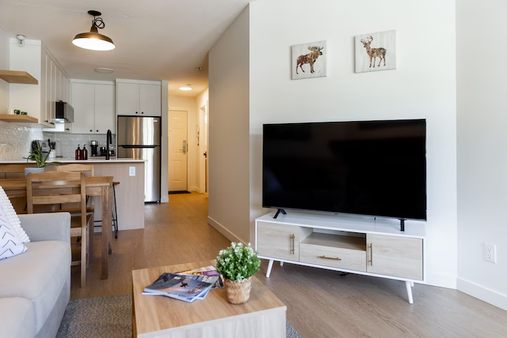 Living room with a modern TV stand, flat-screen TV, and framed animal artwork, adjacent to an open kitchen with dining table, refrigerator, and overhead lighting.