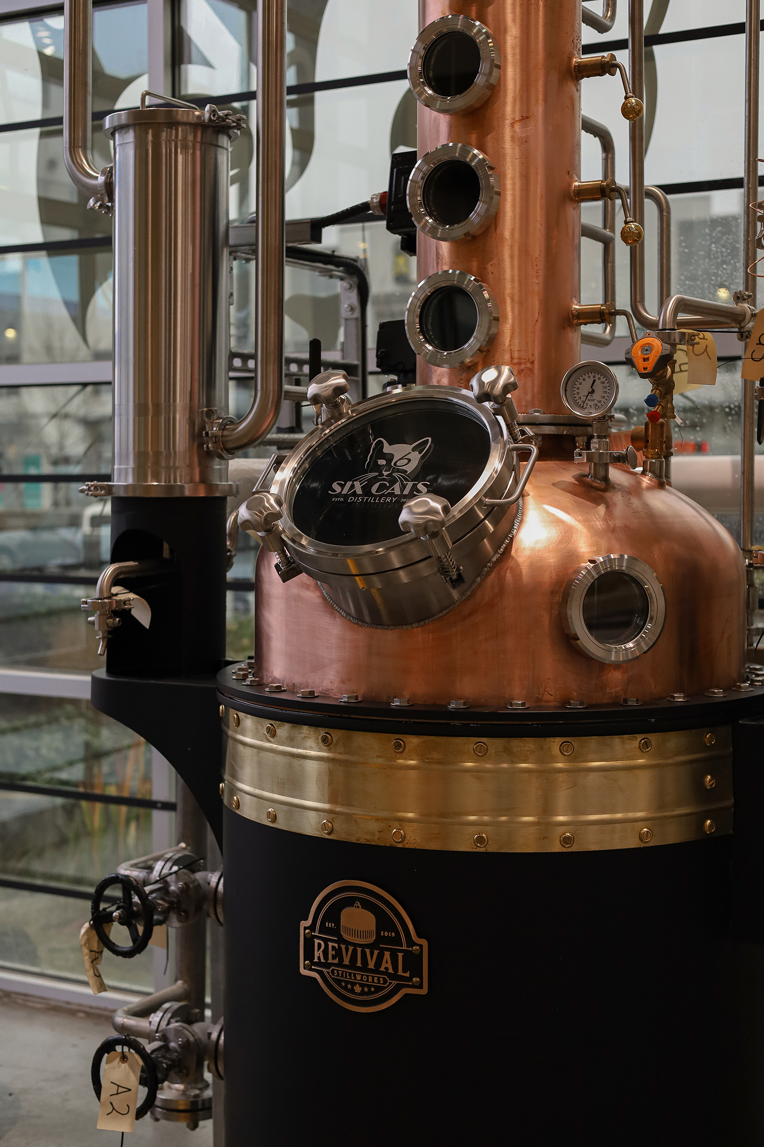 Large copper and steel still used for distilling, with pipes, gauges, and a black sign that says "REVIVAL Stillworks." The setup is inside a building with large windows in the background, with a reflection of trees outside.