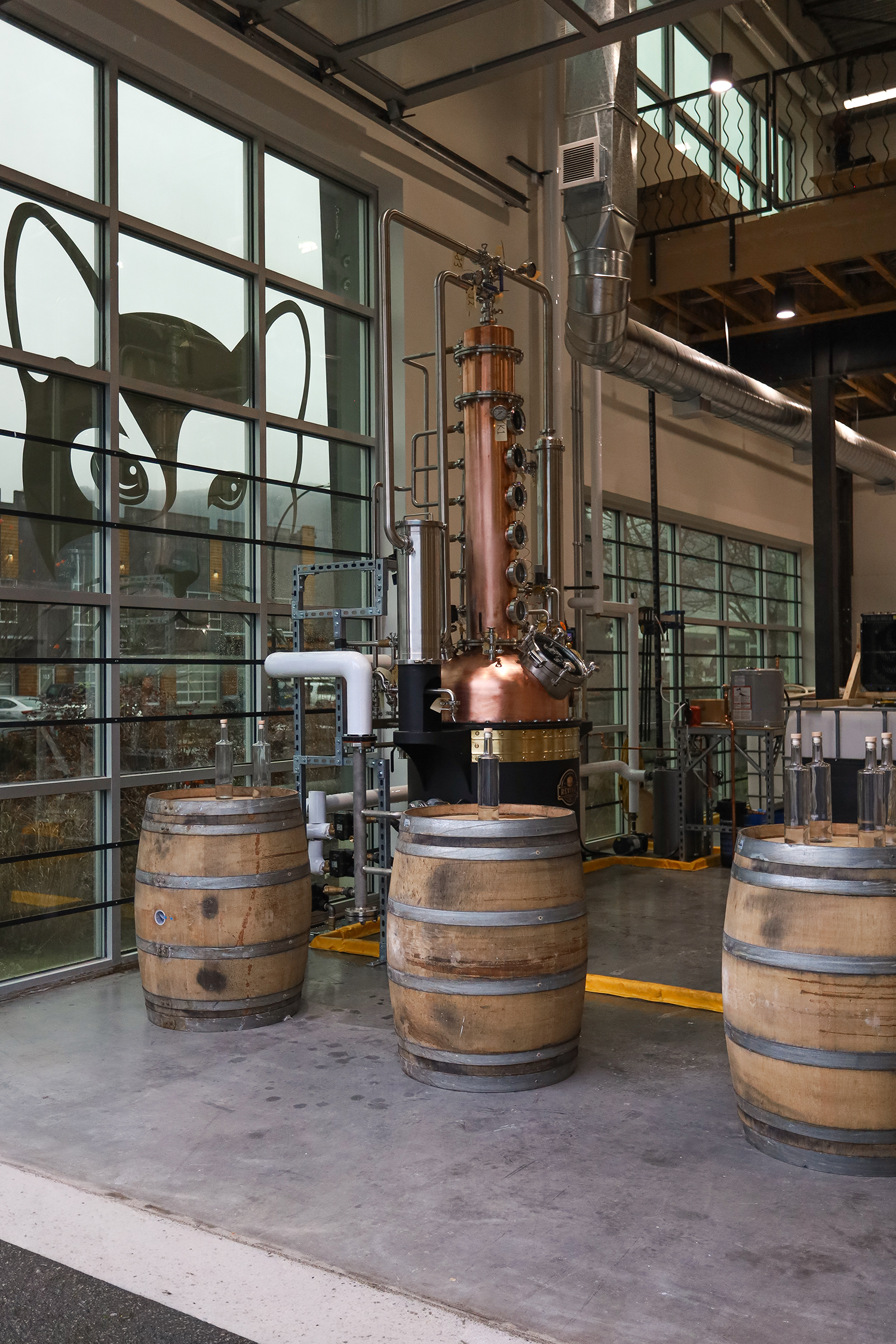 Interior of a distillery with three wooden barrels, industrial distillation equipment, large metal pipes, and a glass wall with a large black cat silhouette painted on it.