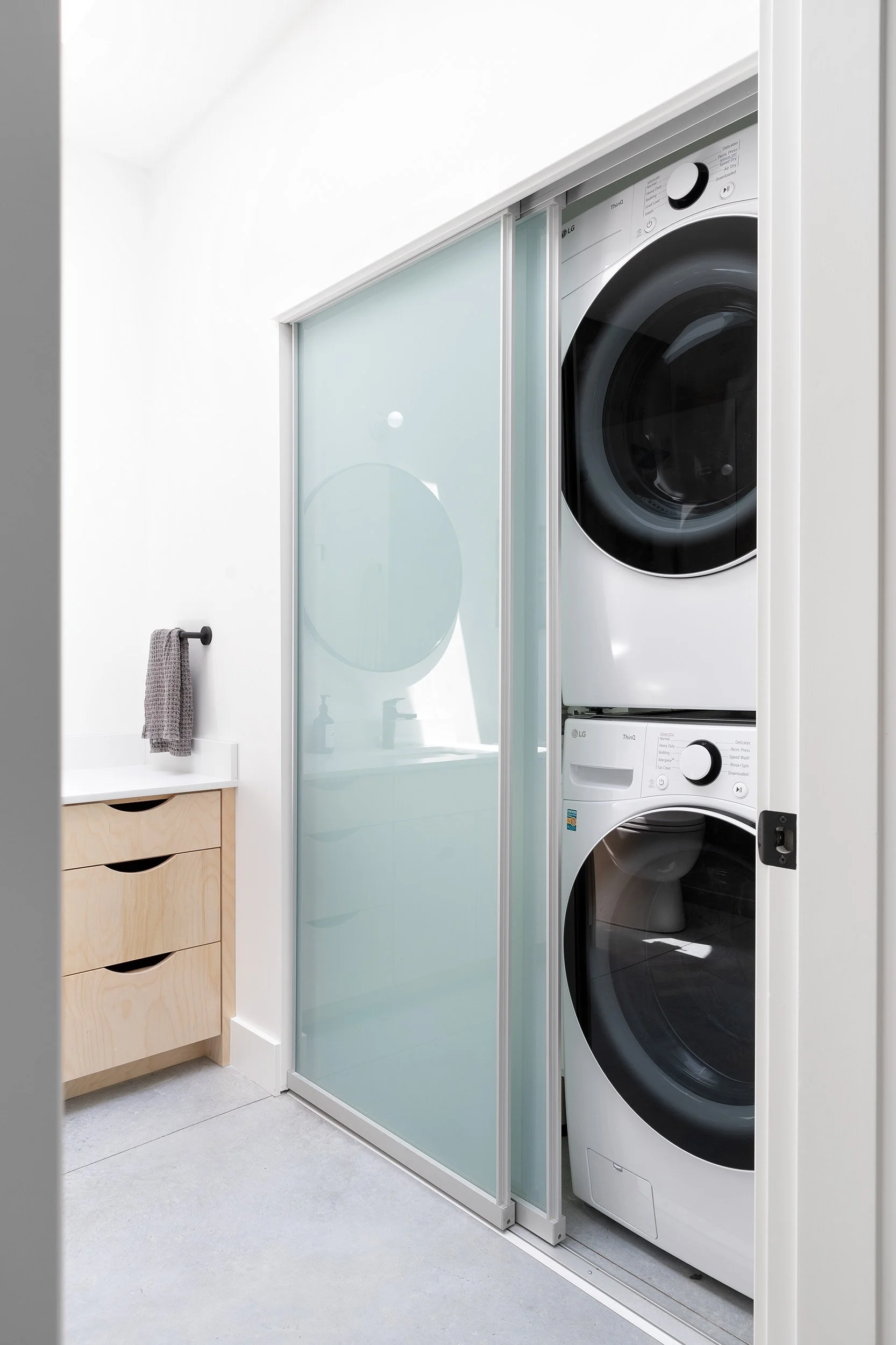A laundry room with a stacked washer and dryer next to a glass door; a wooden cabinet with drawers and a gray towel hanging from a rod.