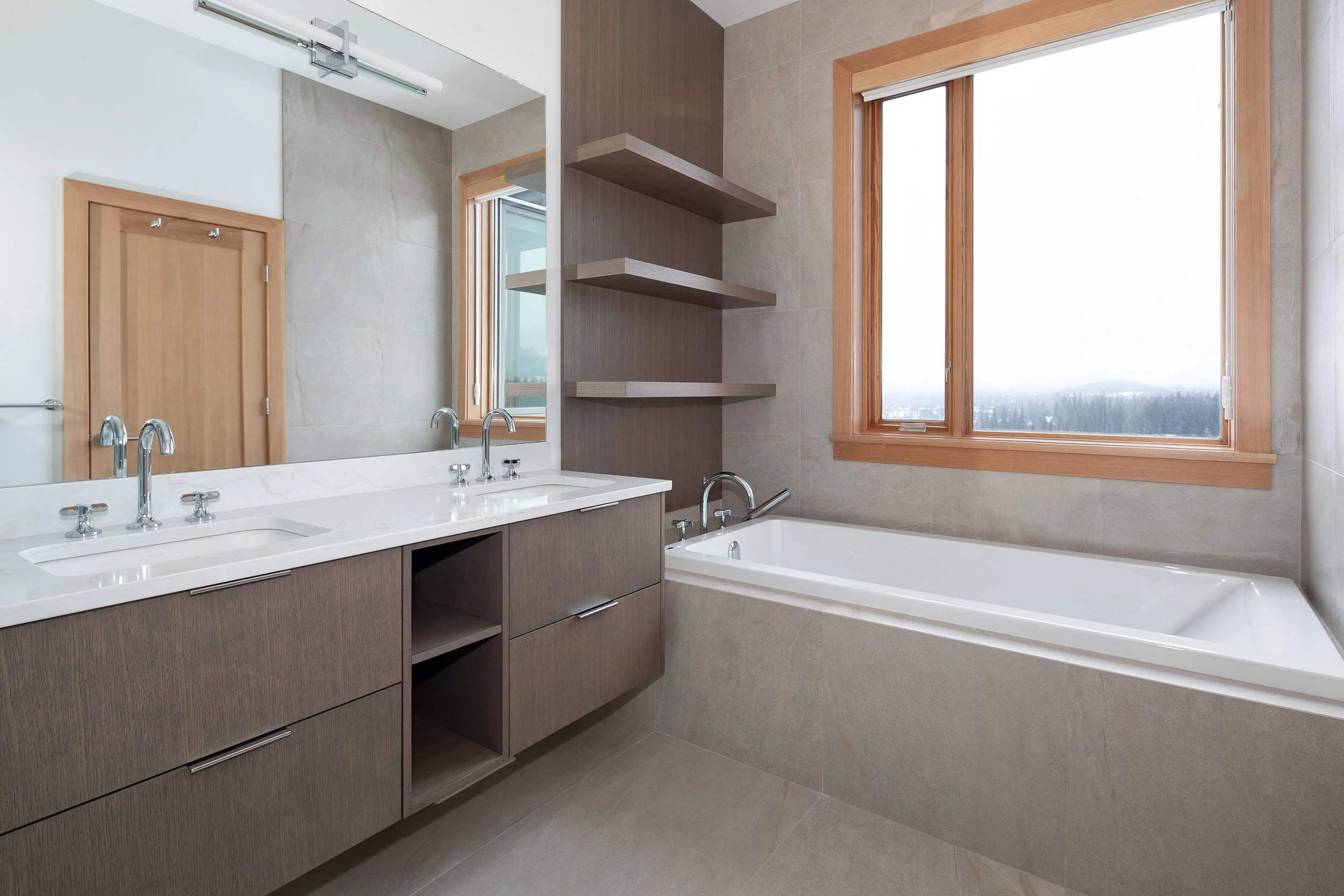 Modern bathroom with double sinks, a bathtub, large window showing a landscape, and open shelving.