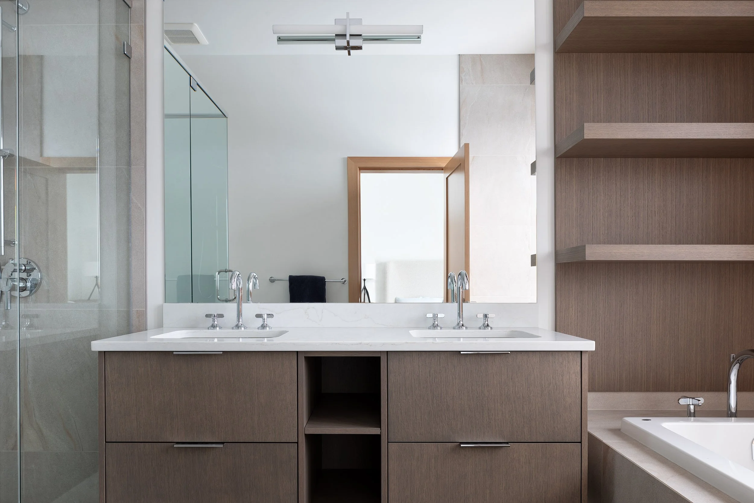 Modern bathroom with a large mirror above a white double sink vanity with wood cabinets, a wooden door with a window, beige tiled walls, a glass shower enclosure on the left, and open wood shelving on the right.