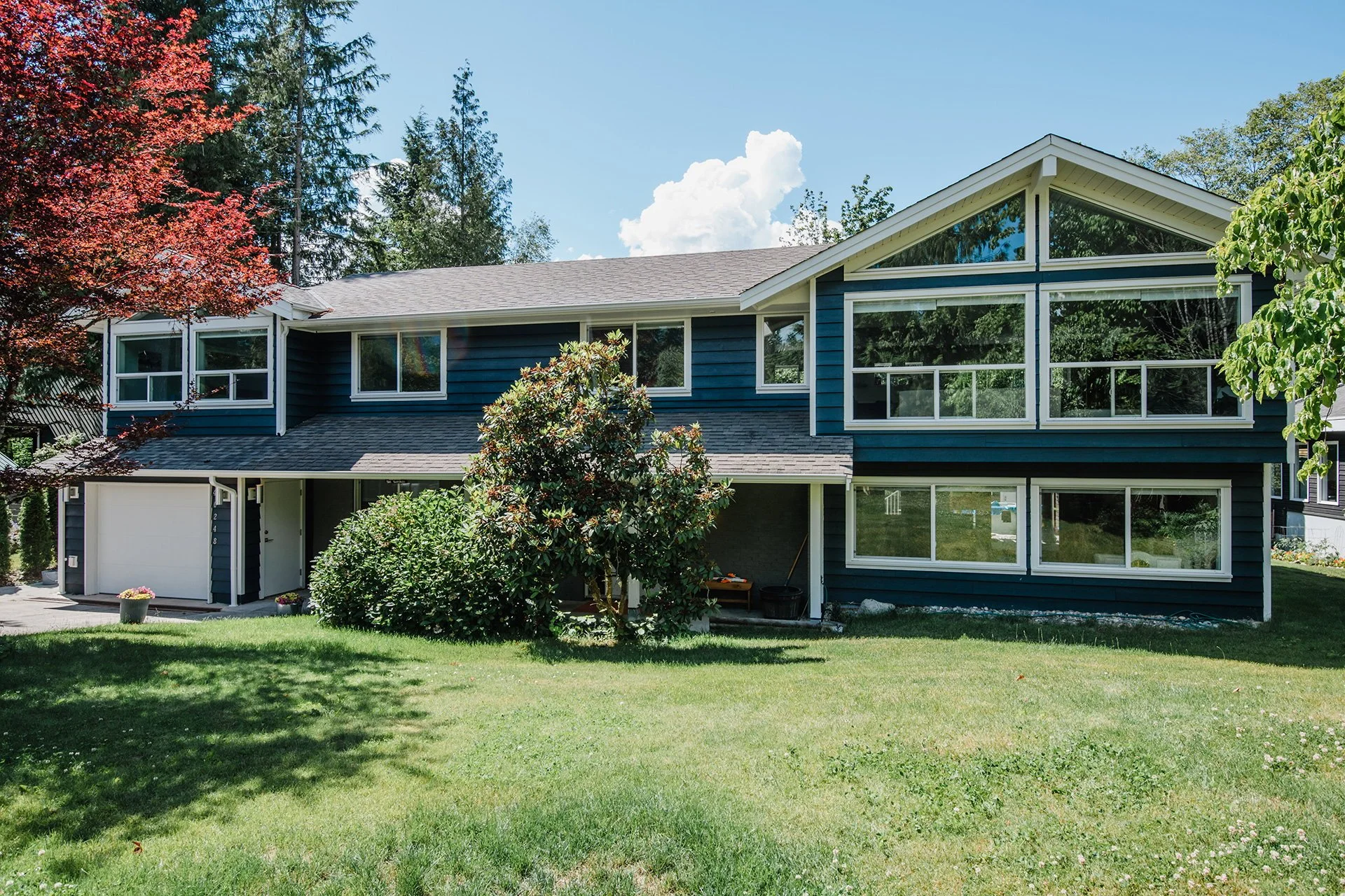 A two-story blue house with large windows, surrounded by green trees and a well-kept lawn under a blue sky with some clouds.