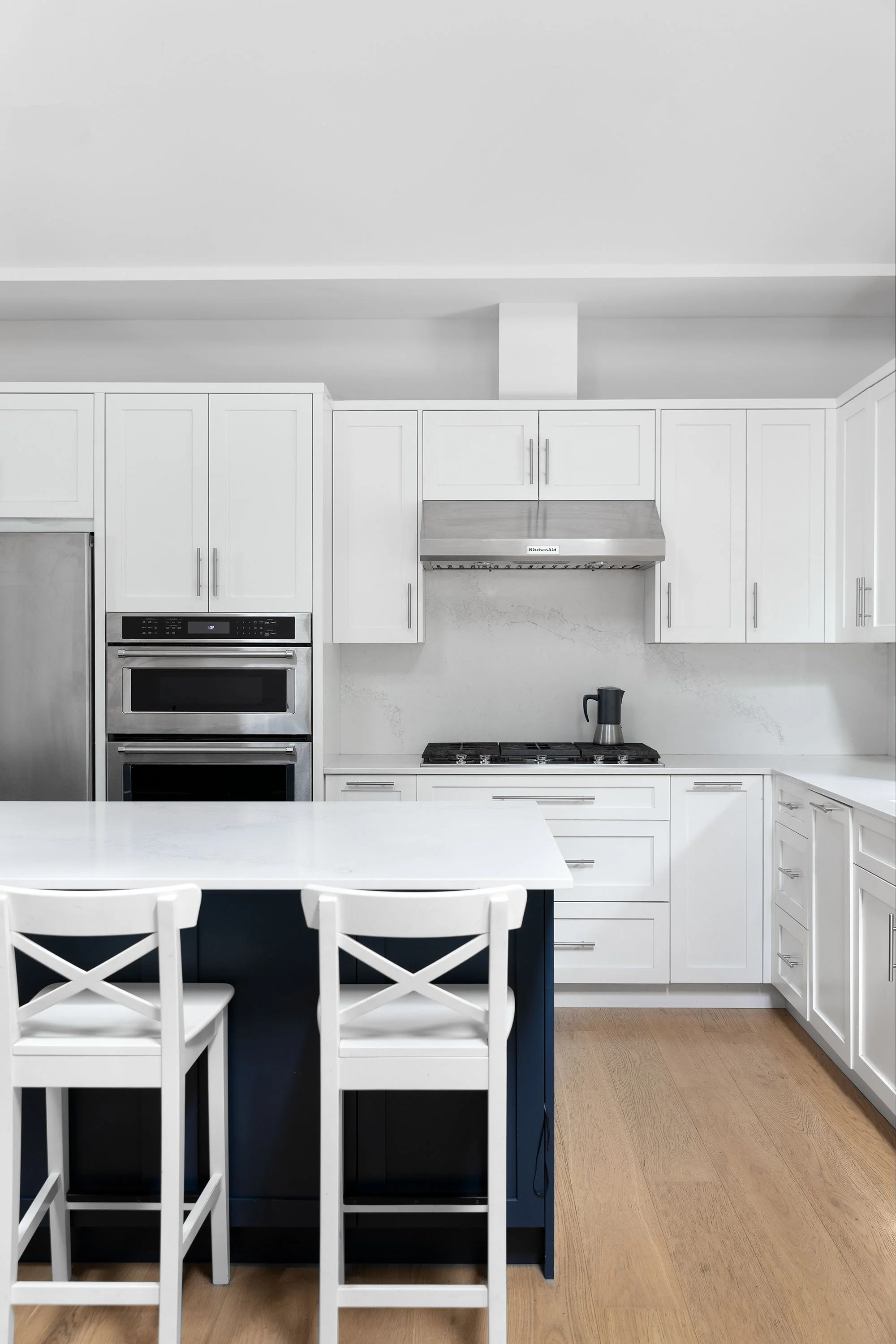 A modern kitchen with white cabinets, a stainless steel oven and range hood, a black kitchen island with white chairs, light wood flooring, and a coffee pot on the countertop.