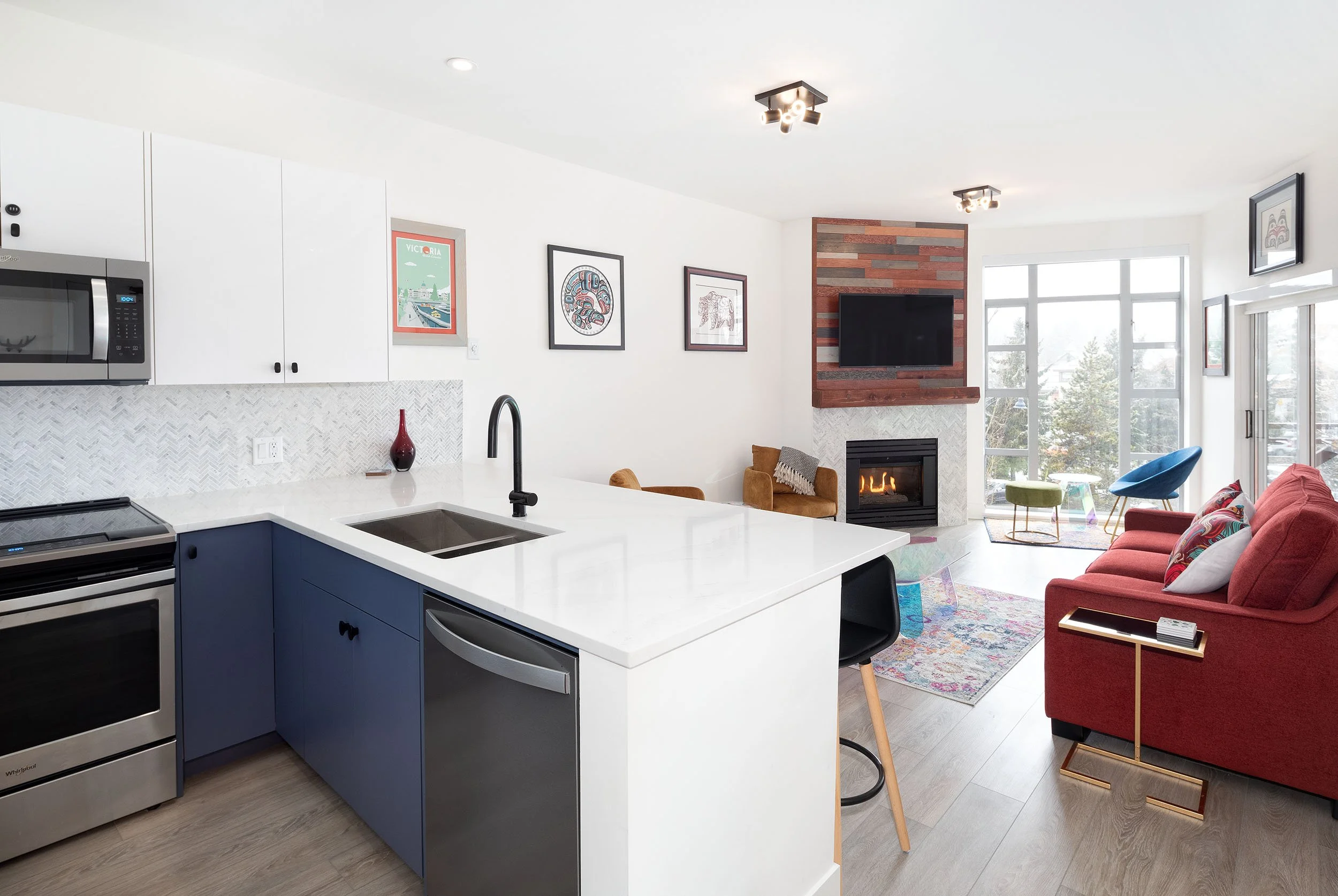 Open-concept living room and kitchen with white walls, modern art on the walls, a fireplace with a wooden accent wall, large windows, and a red sofa with colorful pillows.
