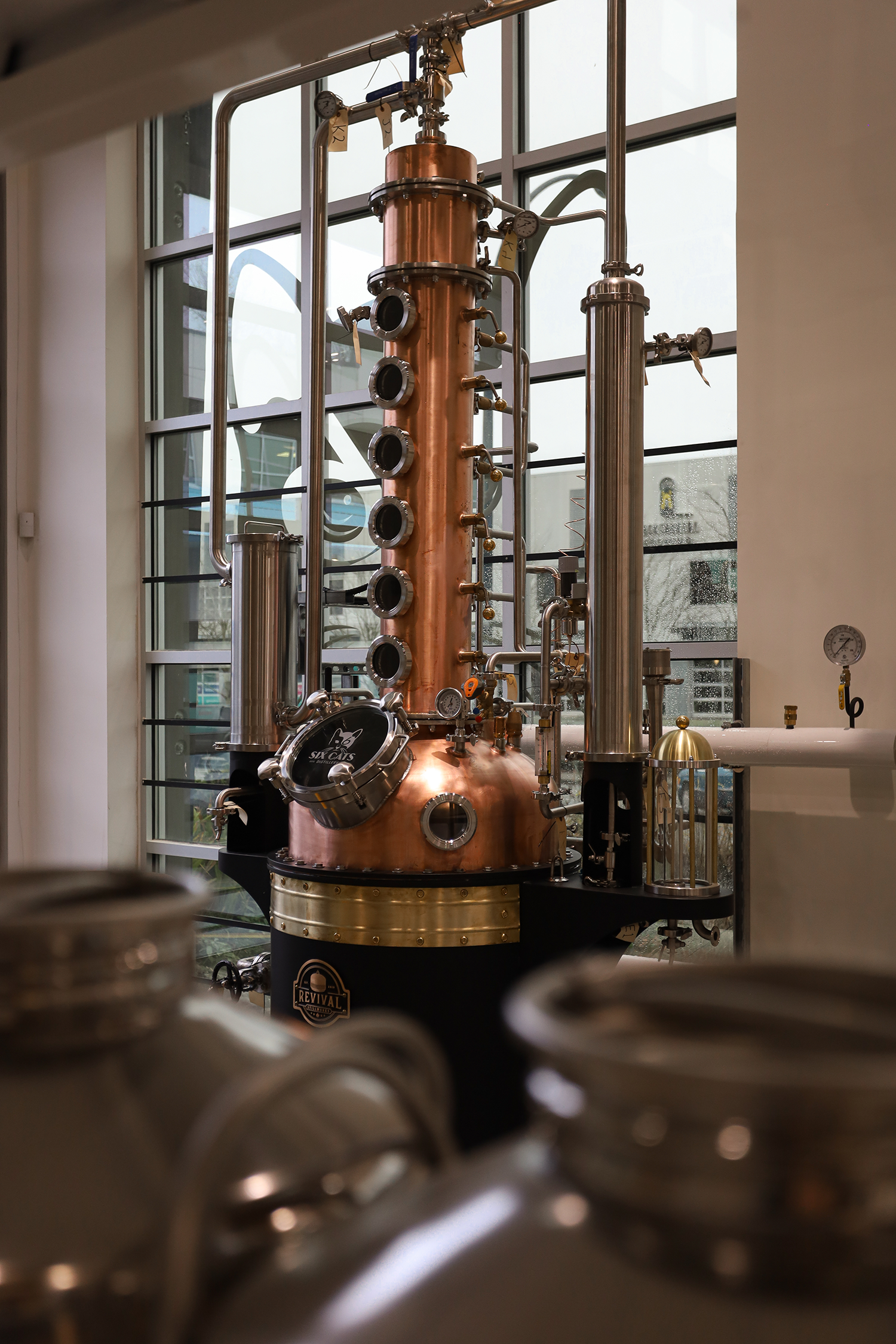 Distillation apparatus with copper and stainless steel components, gauges, and pipes, located inside a building with large windows.