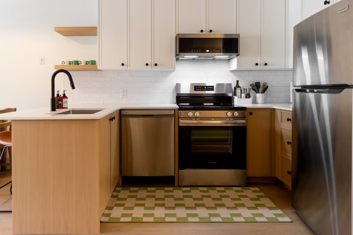 Modern kitchen with white cabinets, stainless steel appliances, a black stove, and a wooden countertop. There is a sink with a black faucet, a fire extinguisher, and a refrigerator, with a patterned rug on the floor.