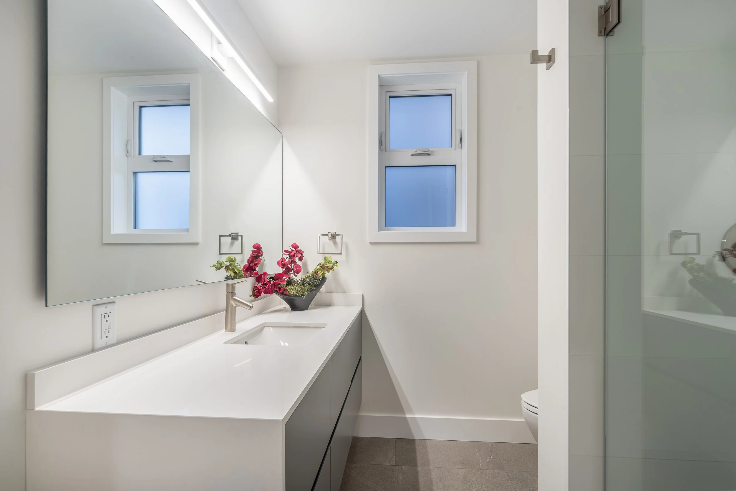 Modern bathroom with a large mirror, two small windows with frosted glass, white countertops, a vessel sink, pink flowers in a black vase, and a glass shower enclosure.