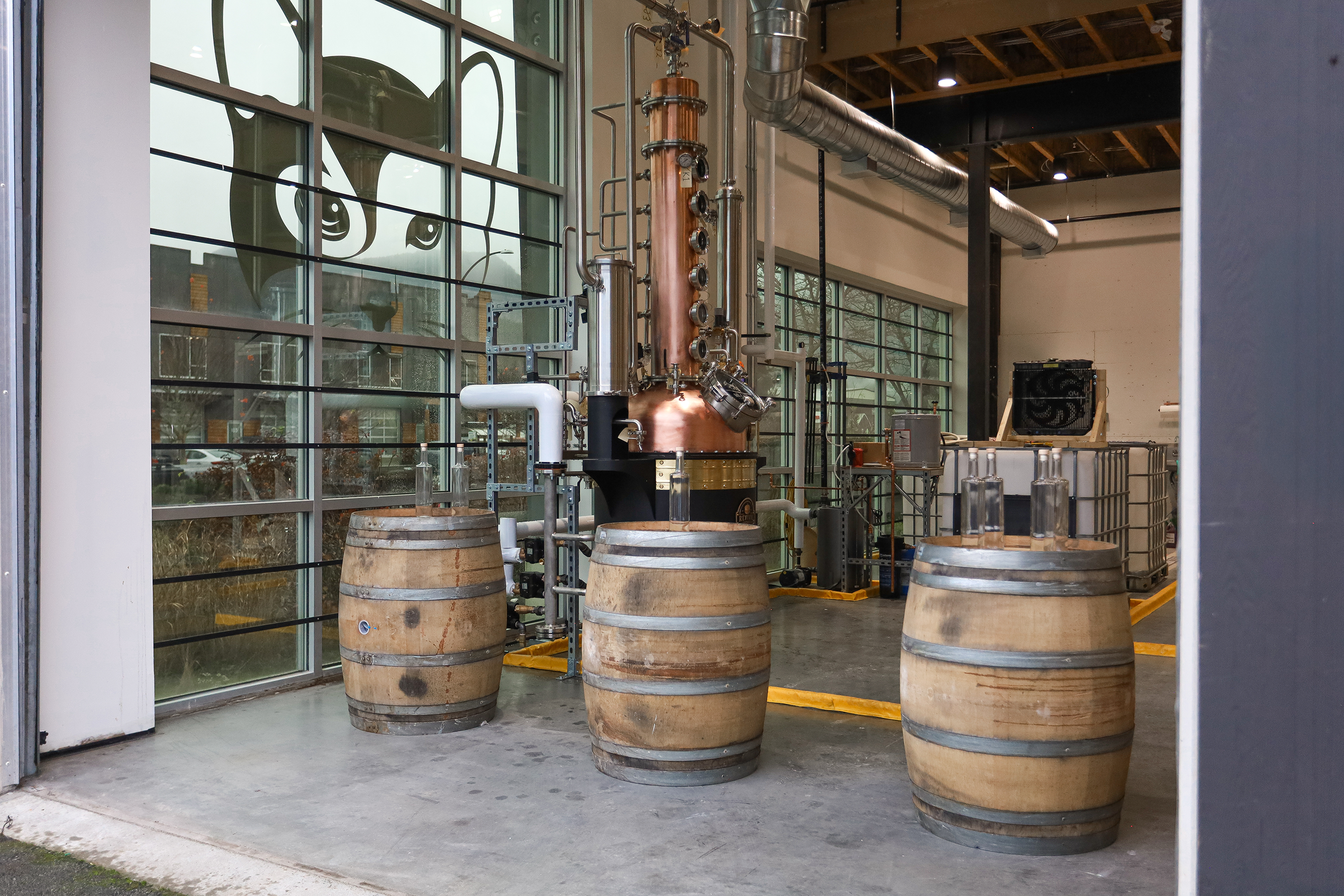 Inside a distillery with four wooden barrels, a copper still, and spray painted artwork of a dog's face on the window.
