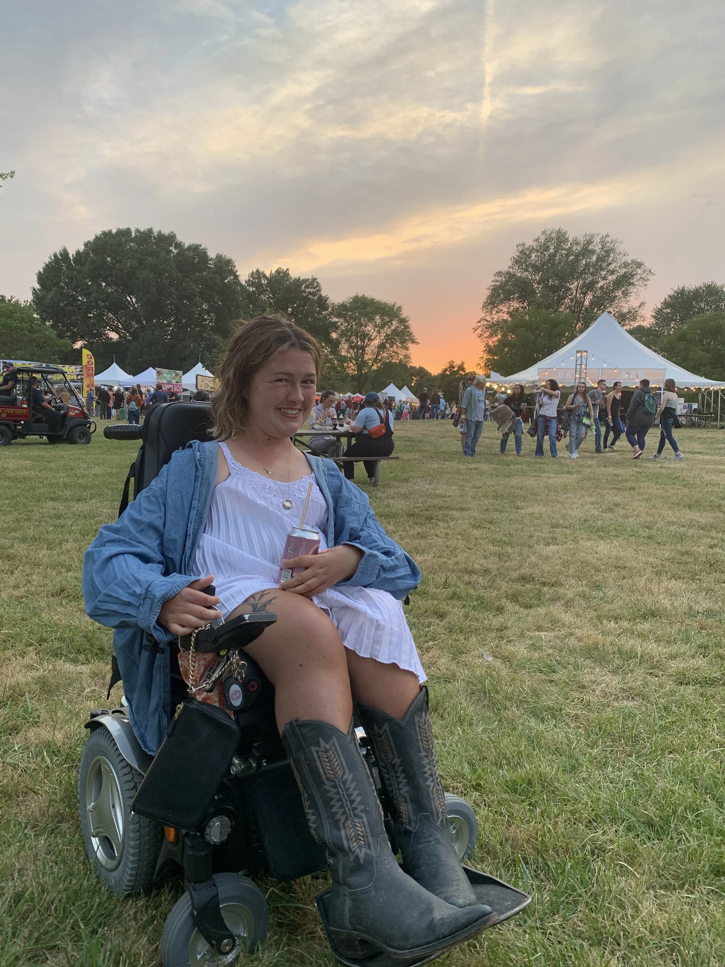 Ellie, a white woman, sits  in a wheelchair in an outdoor music festival. She is wearing cowboy boots and a white dress and holding a drink