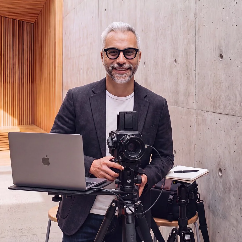 A smiling man with gray hair, glasses, and a beard taking a photograph with a professional camera set on a tripod. He is wearing a gray blazer over a white t-shirt, standing in a modern indoor space with wood and concrete walls, beside a laptop, notepad, and other photography equipment.