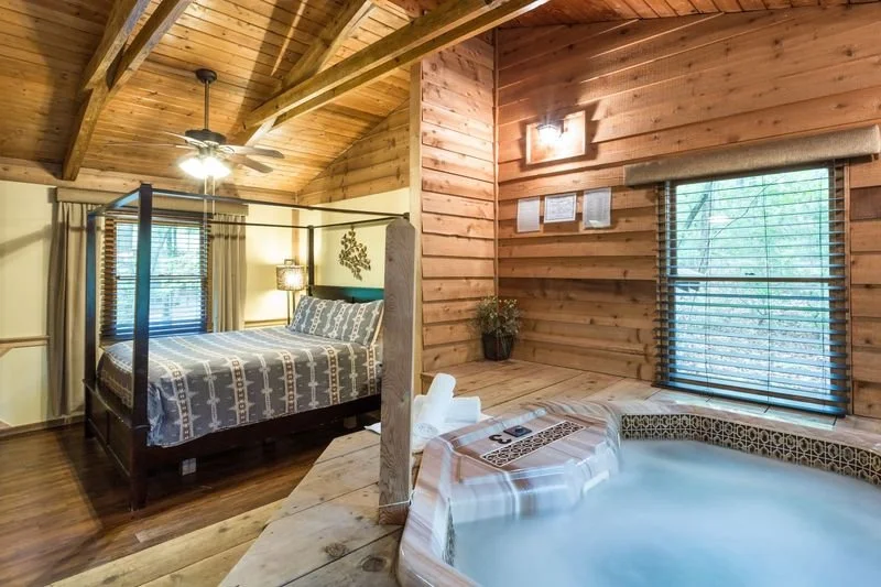 A cozy bedroom with a four-poster bed with patterned bedding, and a corner with a hot tub with towels on the edge. The room has wooden walls and ceiling, with windows and a ceiling fan.