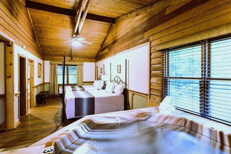 A cozy bedroom with two beds, wooden walls, and ceiling, and a large window with blinds overlooking trees.