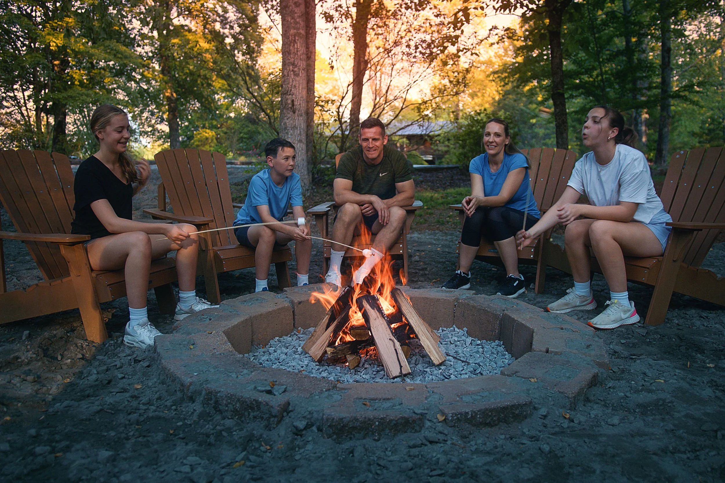 Group of five people sitting around a campfire in a wooded area during sunset, engaged in conversation and roasting marshmallows.