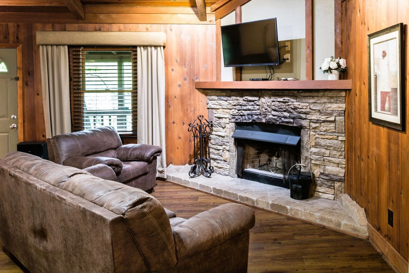 Living room with a stone fireplace, wooden walls, a mounted flat-screen TV, leather sofas, a framed picture on the wall, and a window with curtain and blinds.