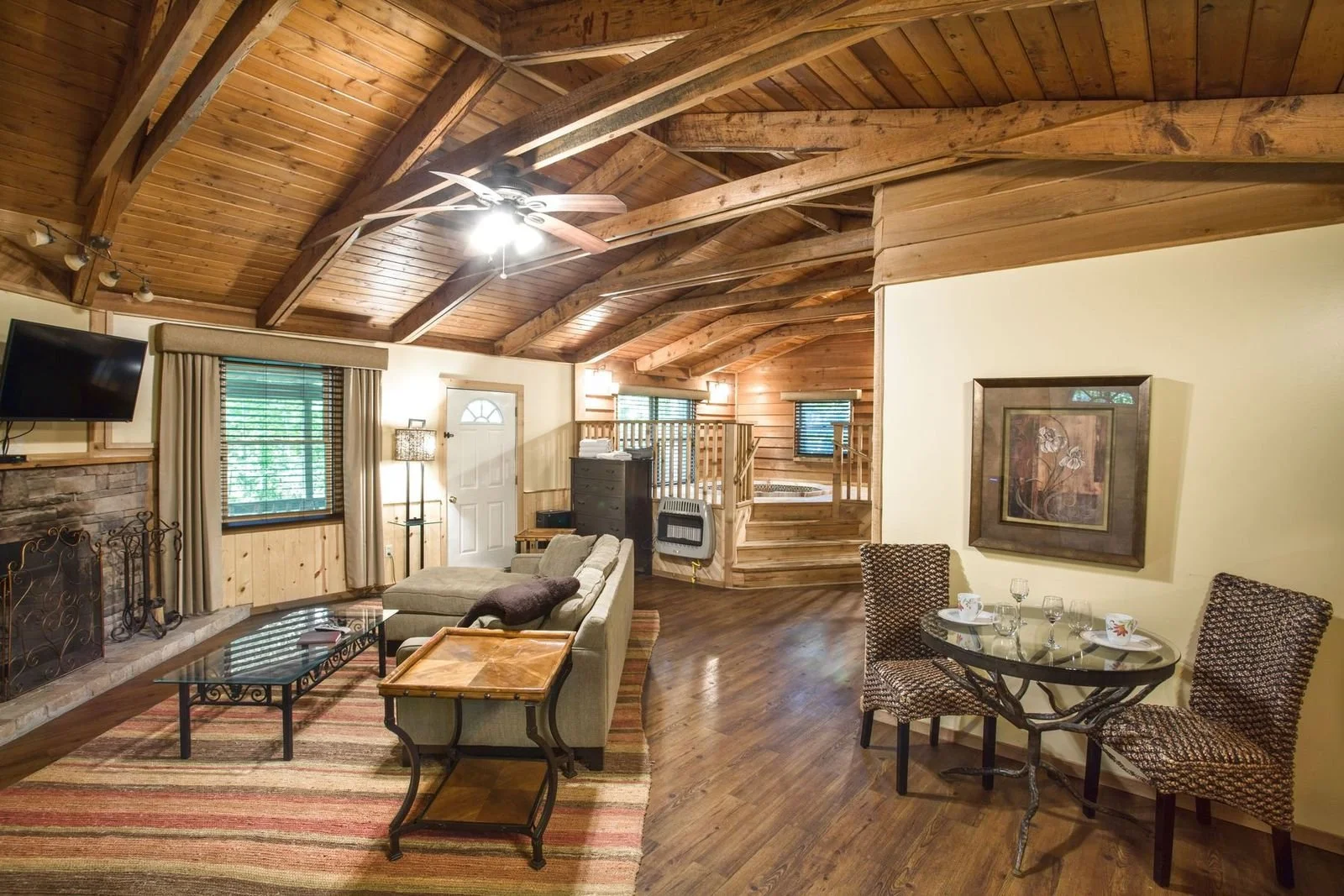 Living room with wooden vaulted ceiling, fireplace, beige sofa, flat-screen TV, dining table with four chairs, and a small staircase leading to a hot tub area.
