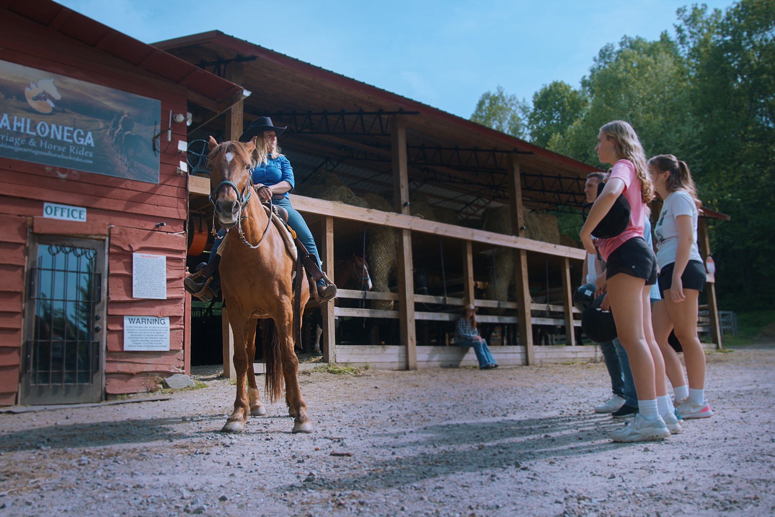 Group of young girls watching a woman riding a horse at a horse riding stable with a sign that reads "Mahonega" and a blue sky overhead.