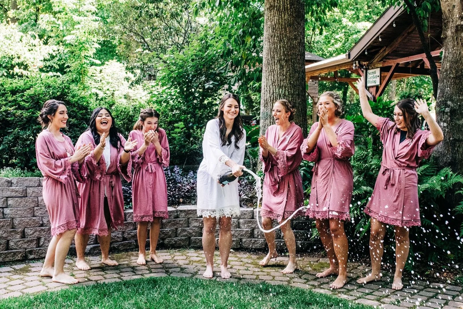 Group of women in pink robes celebrating outdoors, with one woman in white pouring champagne into the air, all smiling and having fun.