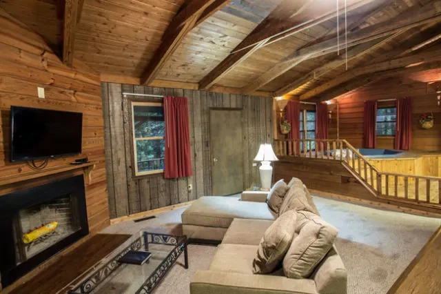 Cozy cabin living room with wooden walls and ceiling, a beige sectional sofa, a glass coffee table, a wall-mounted flat-screen TV, a wood-burning fireplace, a lamp on a side table, windows with red curtains, and a staircase leading to a loft area.