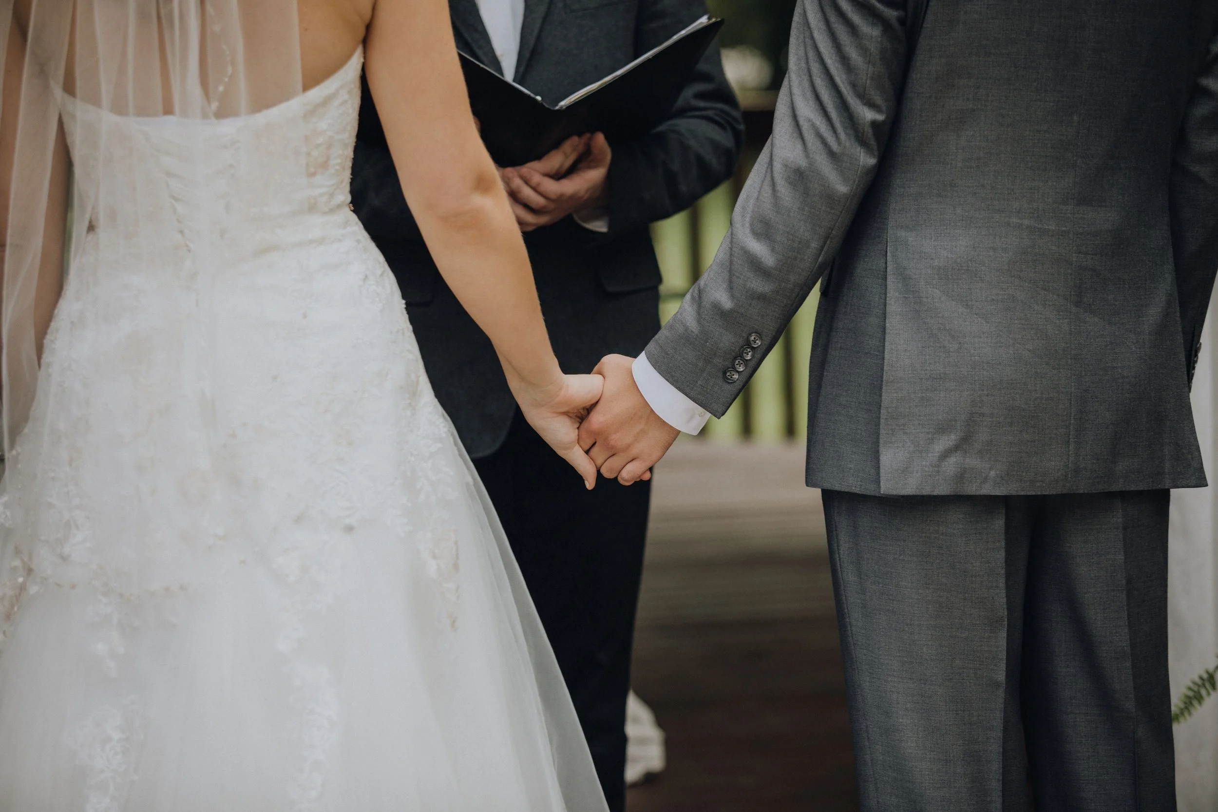 A bride and groom holding hands during their wedding ceremony, with an officiant standing behind them holding a book or folder.