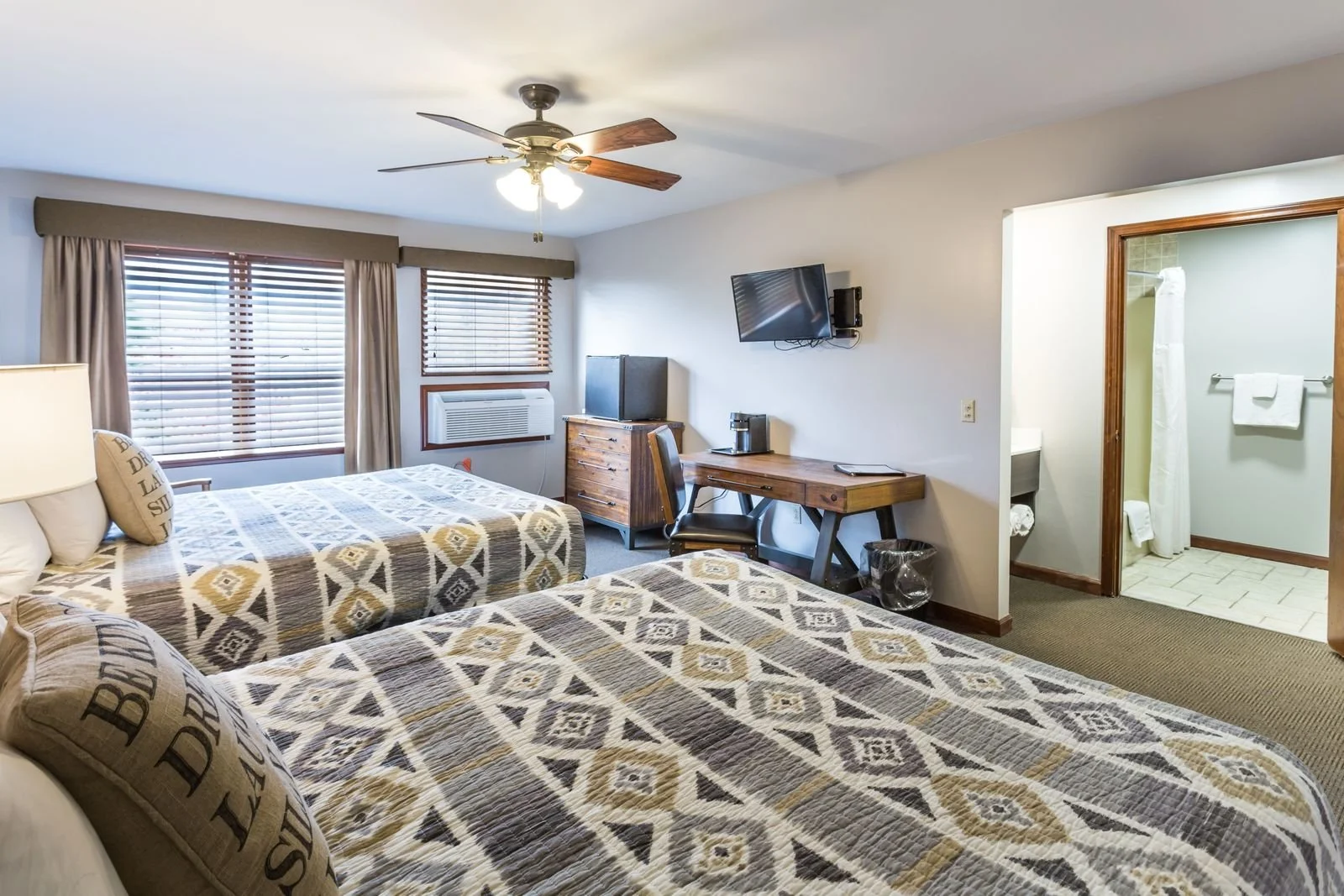 Hotel room with two queen beds, windows with blinds, a ceiling fan, a wall-mounted TV, a wooden desk, and a doorway leading to a bathroom.