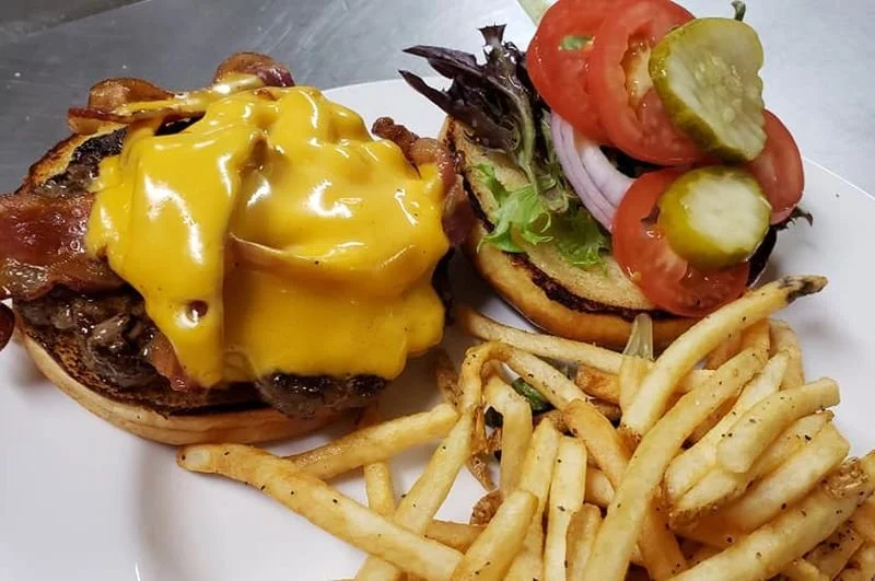 Cheeseburger with melted cheddar, lettuce, tomato, pickles, and onions, served with French fries on a white plate.