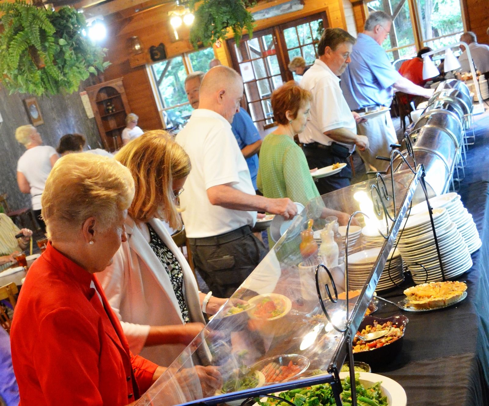 People in line at a buffet in a wooden restaurant, serving themselves food from various dishes including salads, casseroles, and other hot and cold items.