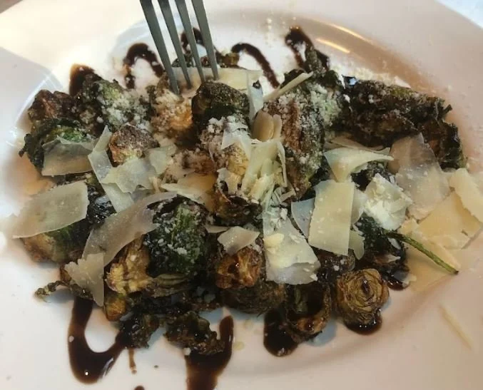 A plate of fried brussels sprouts topped with shaved Parmesan cheese and drizzled with balsamic glaze.