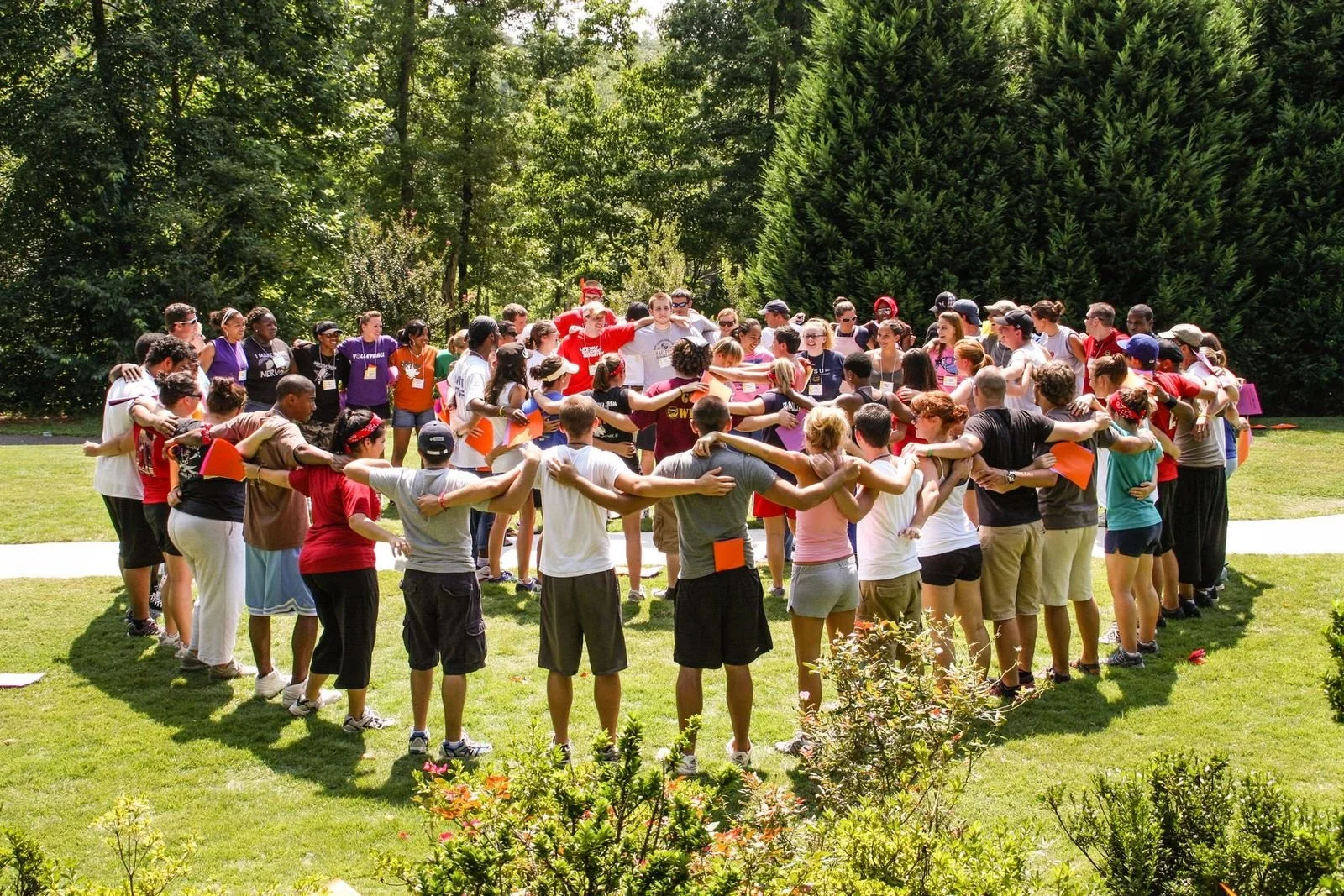 Group of people standing in a circle outdoors on a grassy lawn, holding arms around each other's shoulders, surrounded by trees, participating in an activity or team-building event.