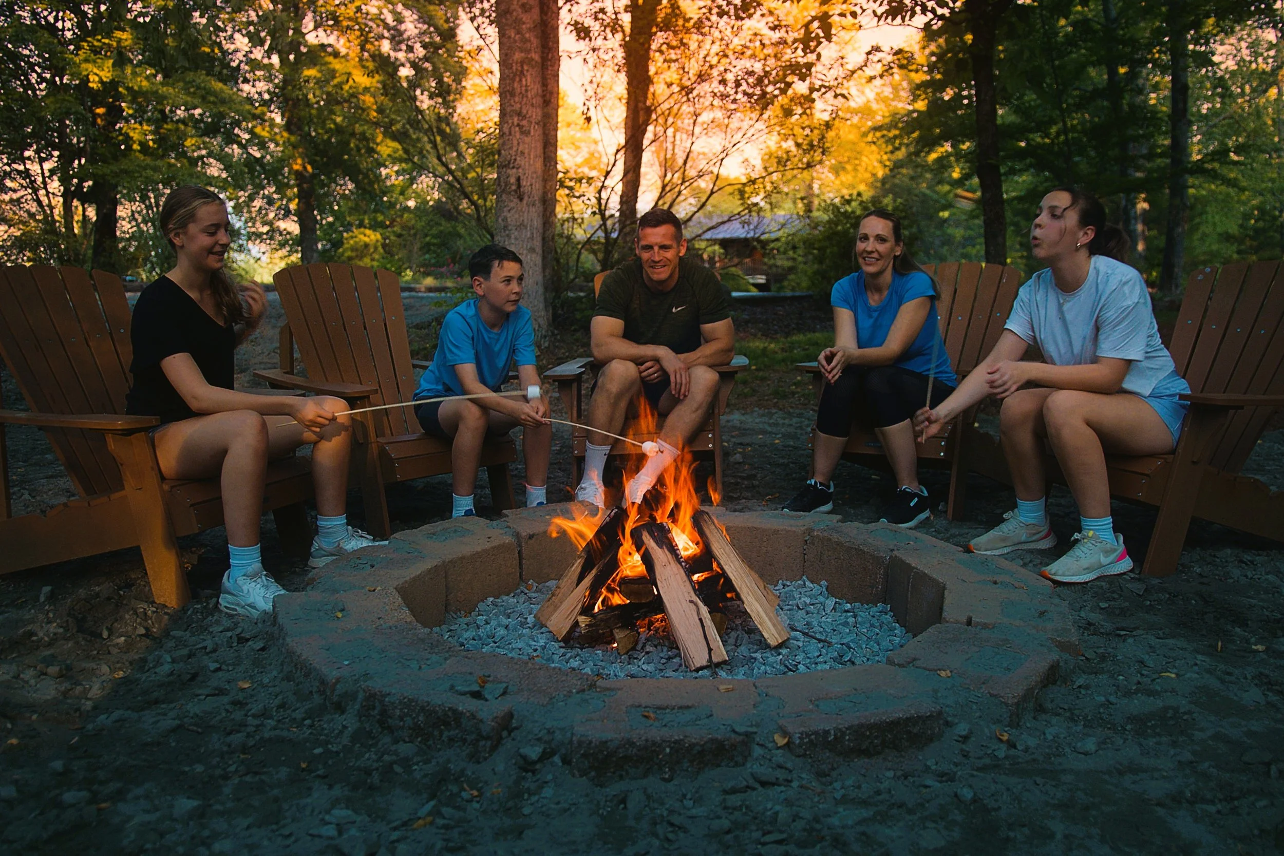 Group of five people sitting around a campfire in a wooded area during sunset, chatting and roasting marshmallows.