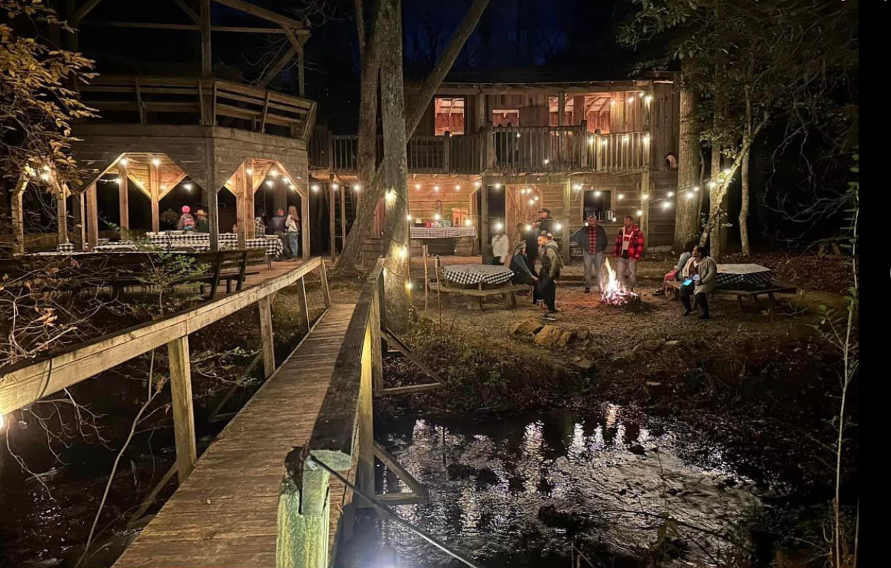Nighttime outdoor gathering at a rustic wooden cabin with string lights, people sitting around a campfire, and a boardwalk leading to the back of the cabin.