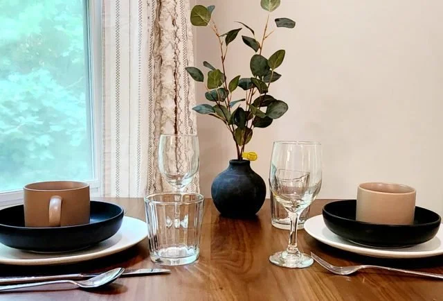 A dining table setup with two place settings, each with a black plate, white dinner plate, beige mug, clear glass, and silverware. A blue vase with green leafy branches is in the center, with a window and curtains in the background.