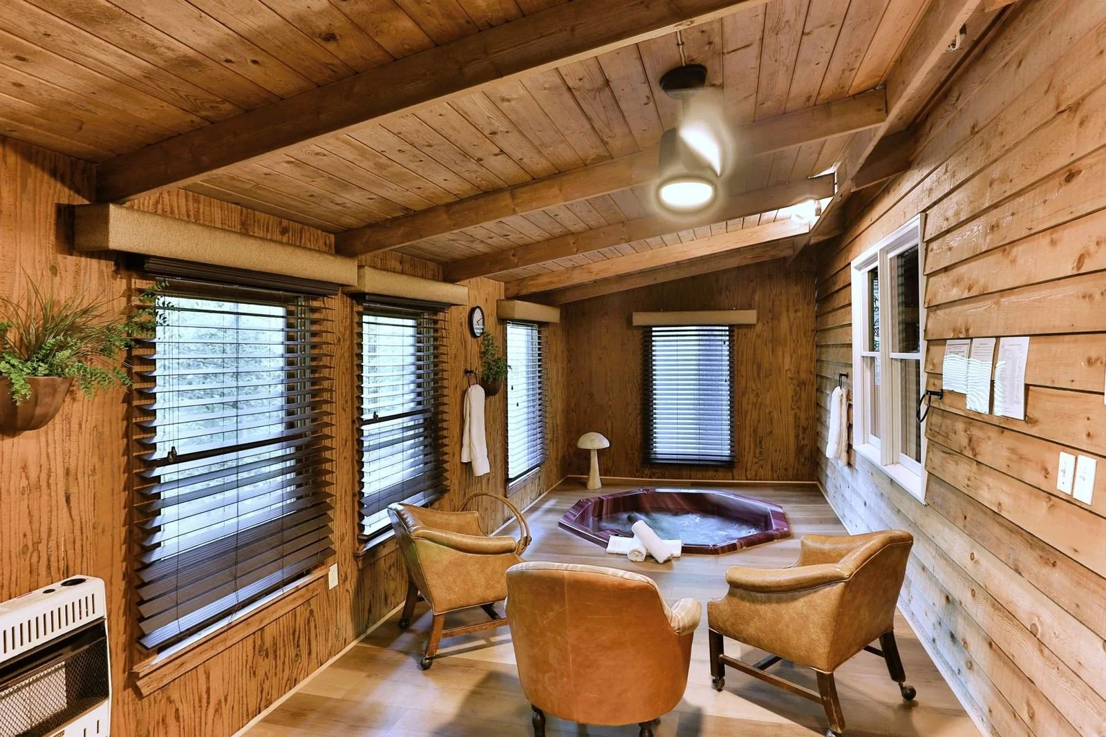 A cozy indoor space with wooden paneling walls and ceiling, featuring a hot tub in the corner, surrounded by four leather chairs, with blinds on the windows and a small lamp on a stand.
