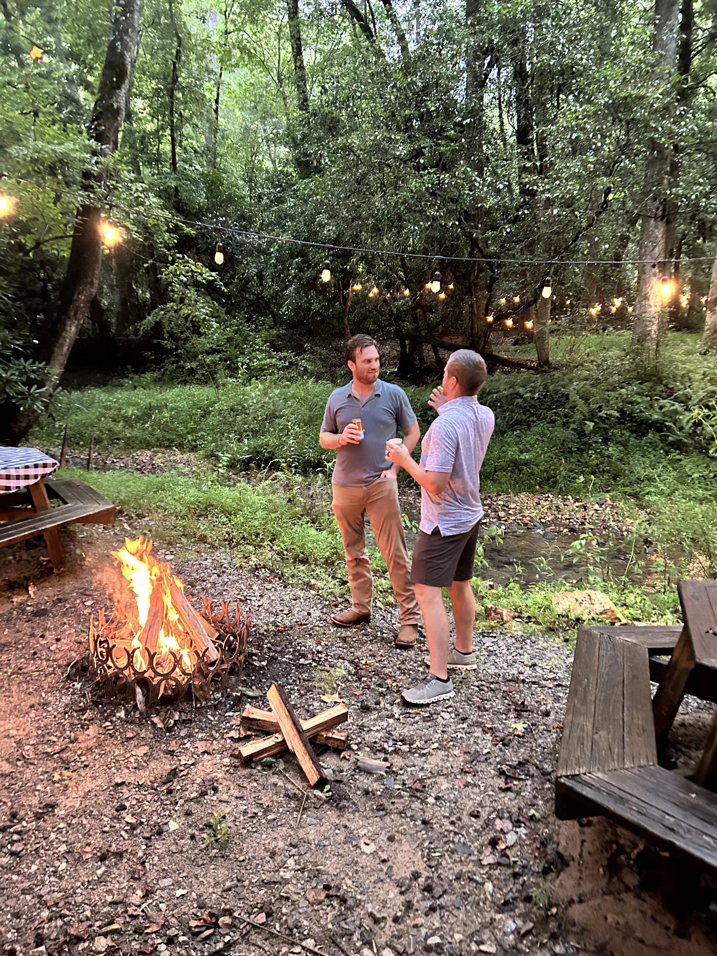 Two men standing by a campfire in a wooded outdoor setting, with string lights hanging above them and surrounded by greenery.