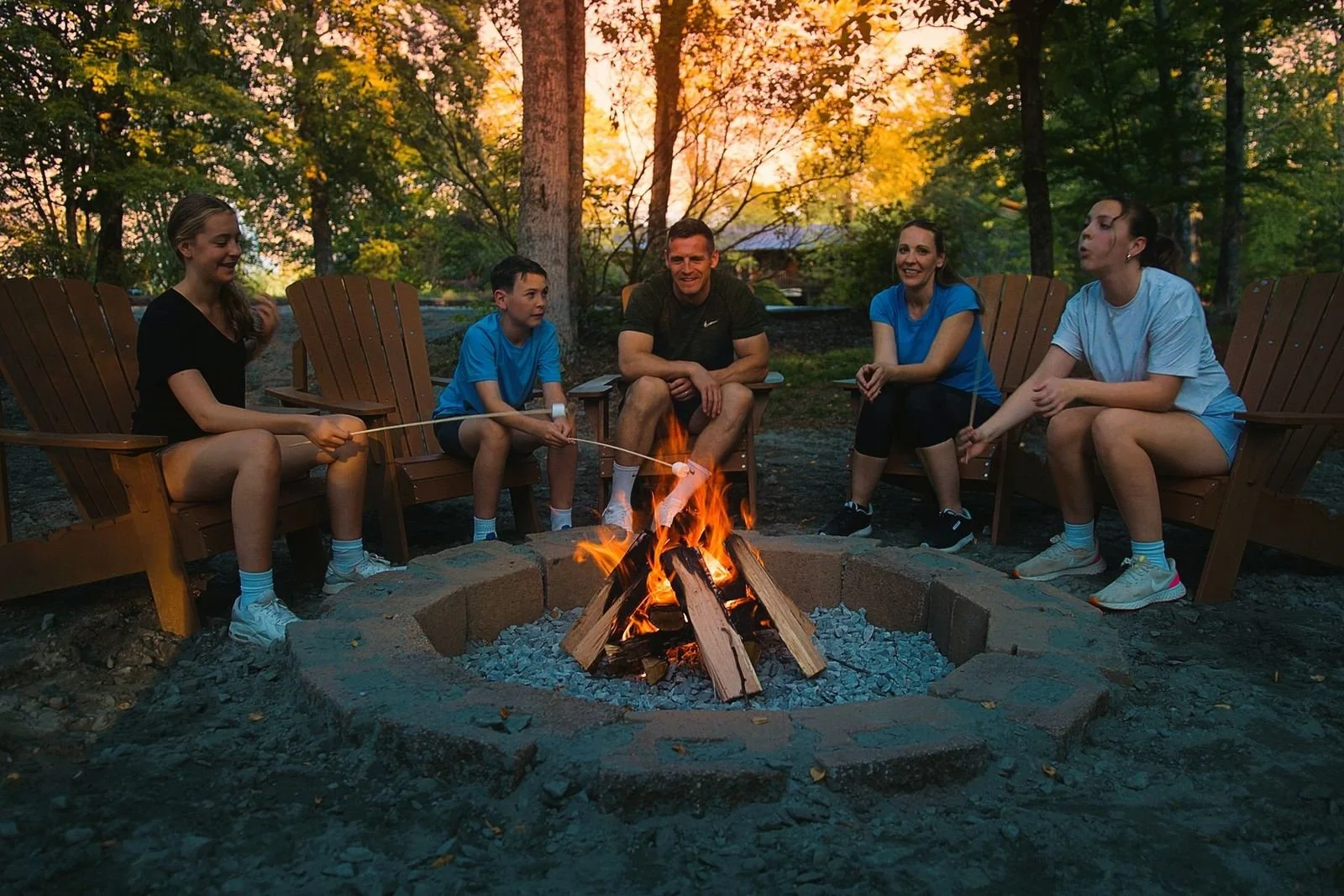 A group of five people sitting around a campfire in an outdoor setting during sunset, with trees and a wooded area in the background.