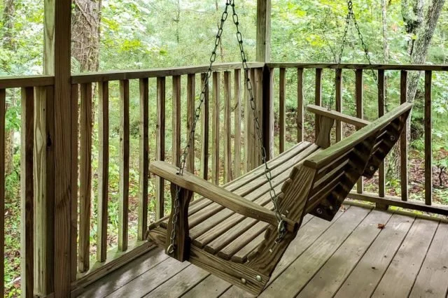A wooden porch swing hanging from a wooden deck railing on a porch surrounded by trees.