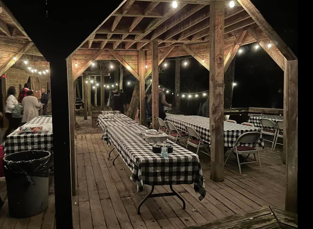 Nighttime outdoor area with a wooden structure and string lights, long tables with checkered tablecloths, chairs, and some people standing and talking.