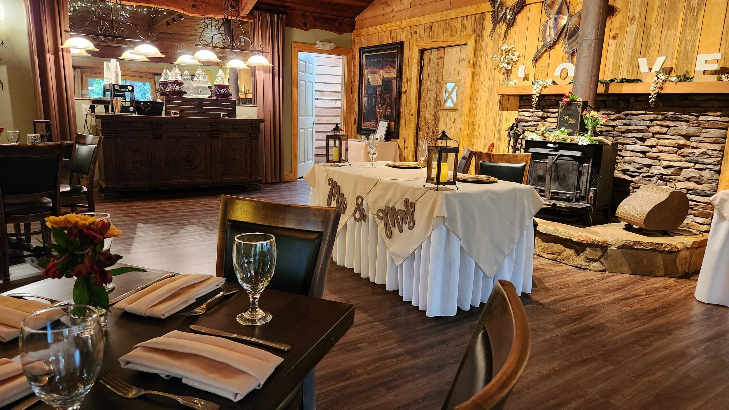 A cozy, rustic dining room decorated for a celebration with a table set with napkins, glasses, and silverware, adorned with a flower arrangement. There is a fireplace with decorative flowers and a large sign spelling 'LOVE' on a wooden wall. The room