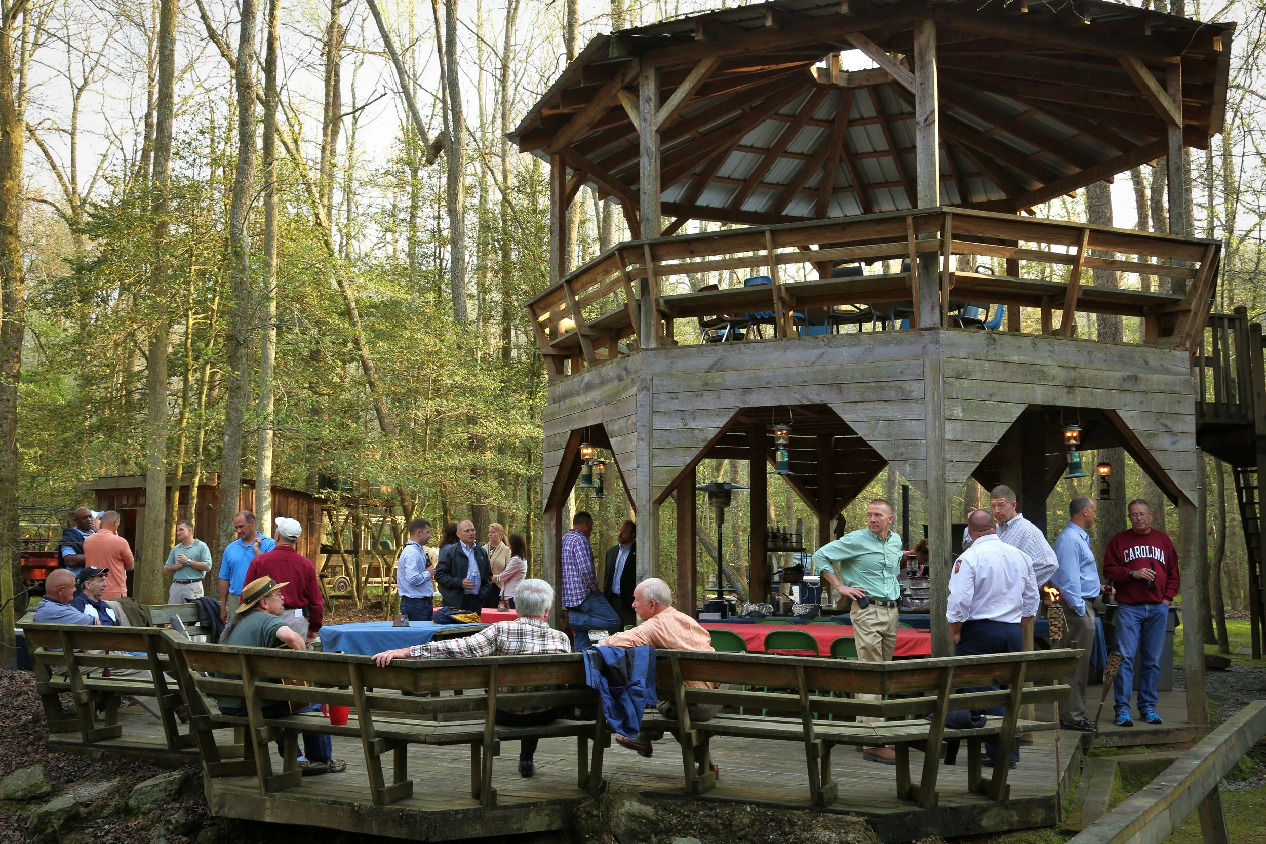 People gathering outdoors at a wooded area with a two-story wooden gazebo. Some people are sitting on benches, others are standing and talking, with tables covered in red or checkered tablecloths. The scene suggests a social event or picnic.