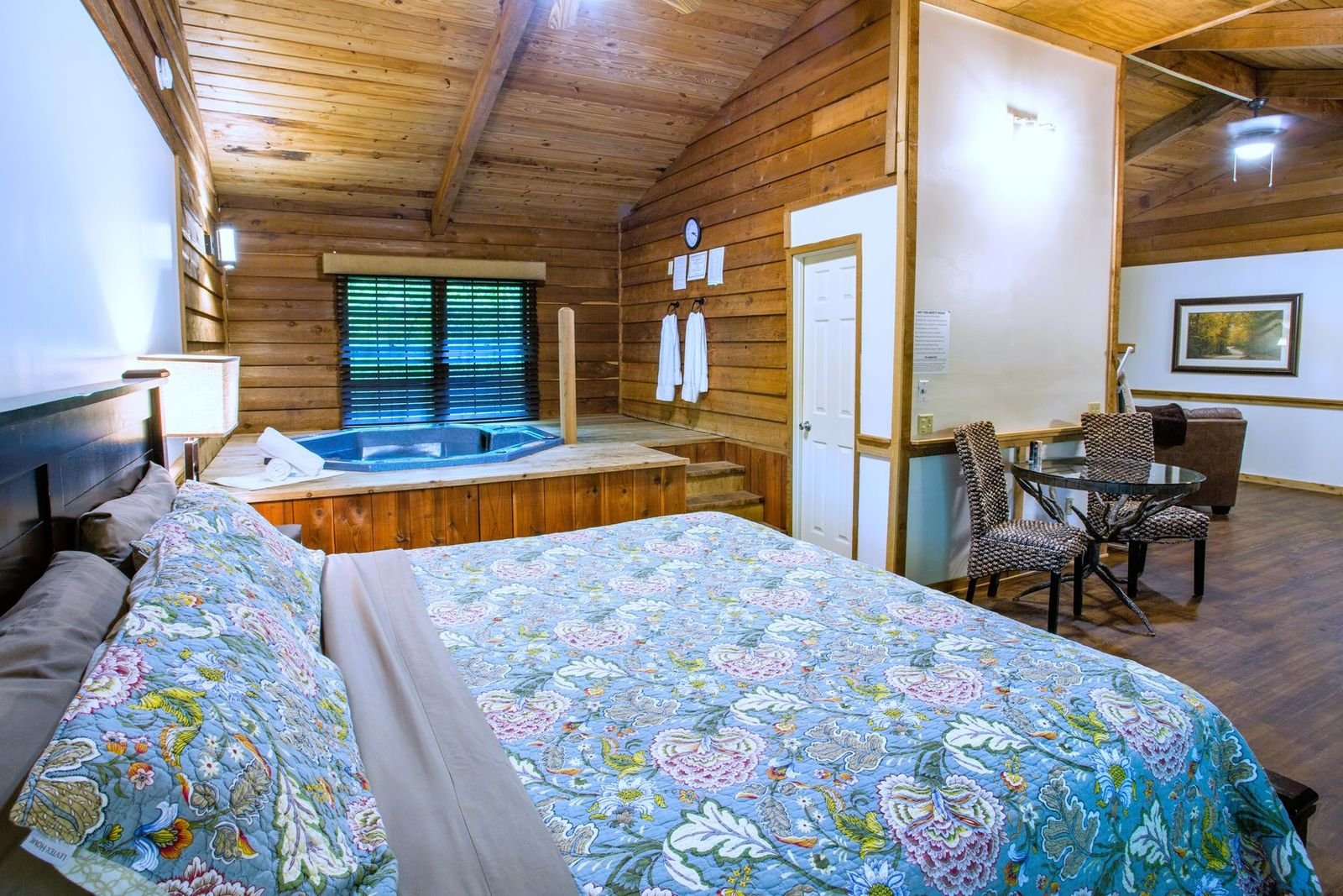 A cozy hotel room with a large bed covered in a floral quilt, wood-paneled walls, and a hot tub near a window with blinds. There are two white bathrobes hanging on the wall, and a door next to the hot tub. In the background, there is a seating area w