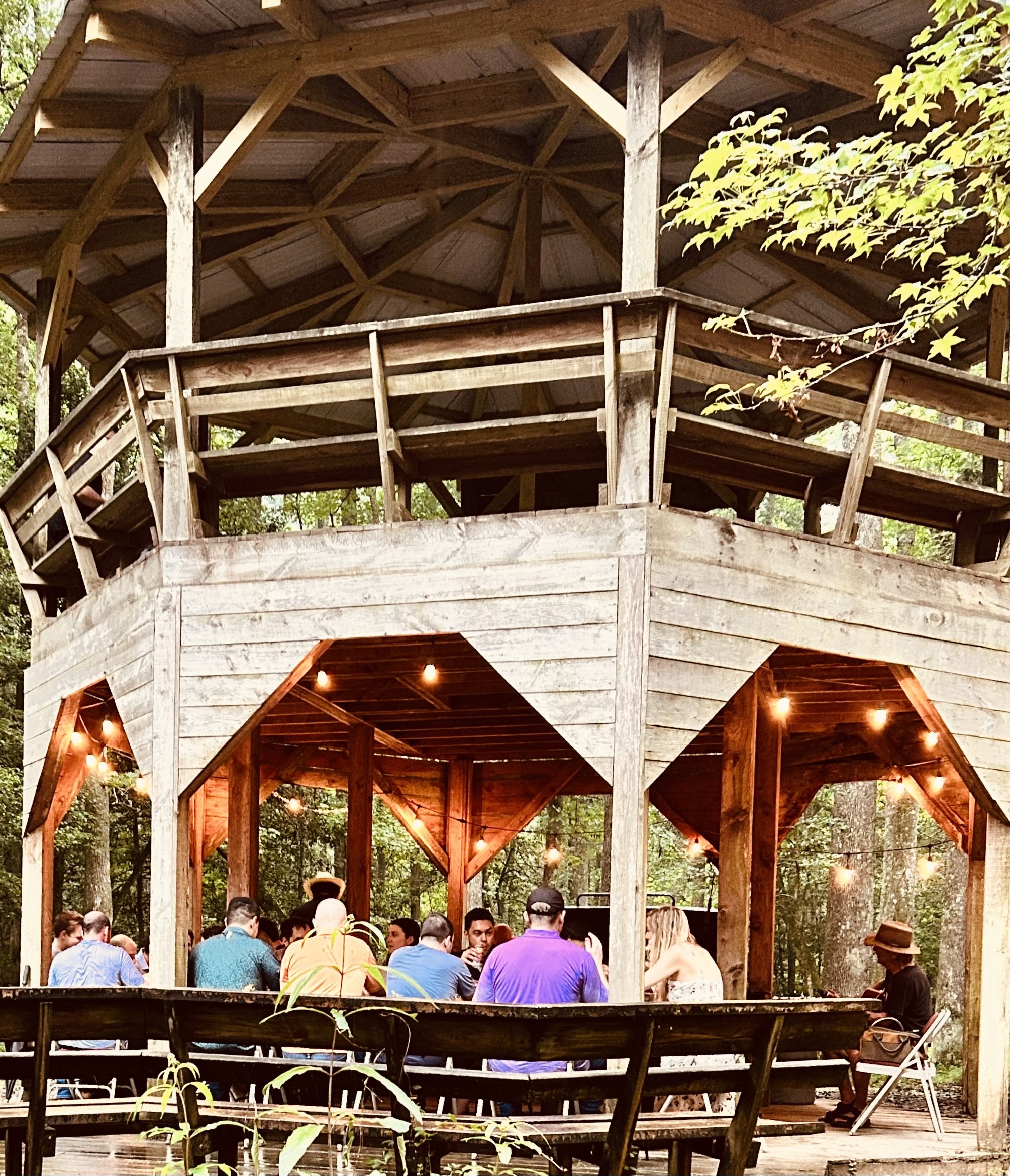 Wooden outdoor stage with people gathered underneath, surrounded by trees and string lights, in an outdoor setting.