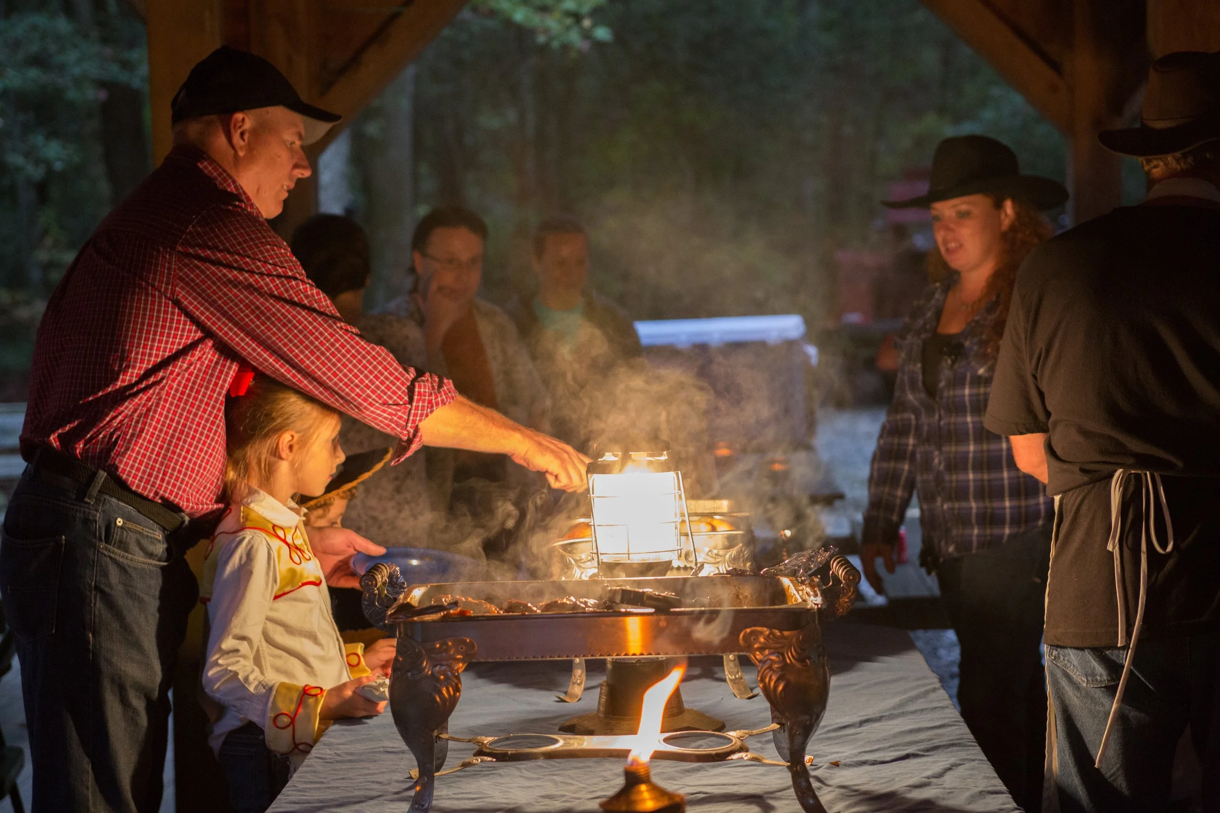 People gathered around a grill with flames, roasting marshmallows at night outdoors, under a wooden shelter.