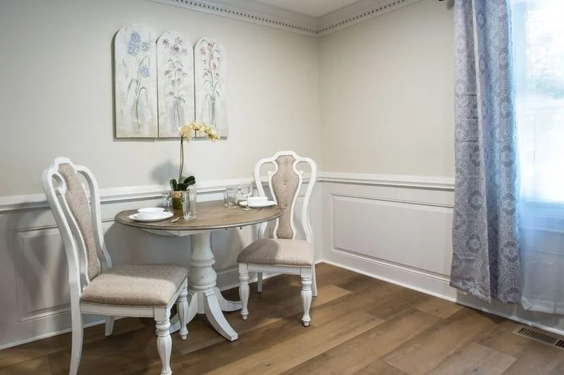 A small round dining table set for two with plates, glasses, and a flower vase, in a room with cream-colored walls, floral wall art, wooden flooring, and a window with patterned curtains.