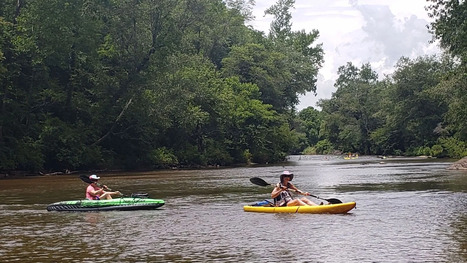 People kayaking on a river surrounded by lush green trees under partly cloudy sky.