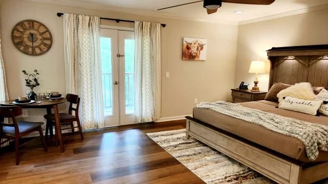 A spacious bedroom with a large bed, nightstand with a lamp, a door leading outside, a small dining table with chairs, a large wall clock, and a window with curtains.